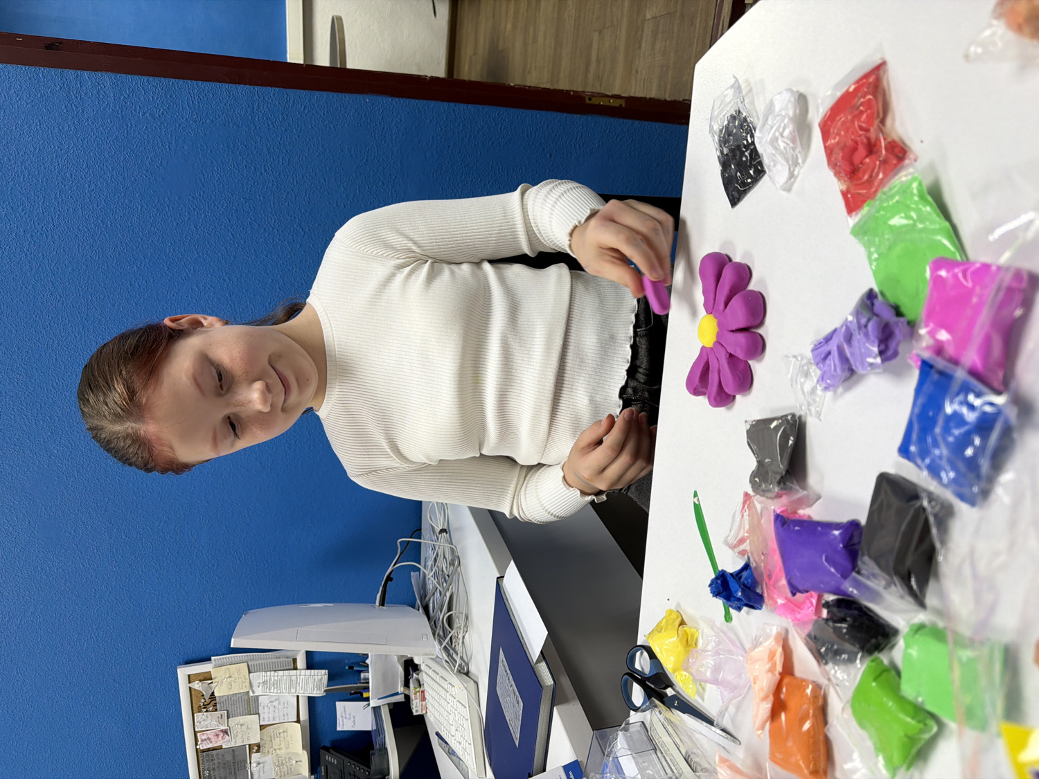 A young person, likely a girl with light brown hair and a white long-sleeved, ribbed top, is engaged in a crafting activity indoors in Chaikovsky, Russia. She is seated at a desk, focused on shaping colorful clay or play-doh on a large white surface.
The main activity involves working with various colors of clay, which are presented in small, clear plastic bags arranged around the white surface. Visible colors include purple, yellow, red, green, blue, pink, black, grey, white, orange, and light pink. A prominent object on the white surface is a partially completed purple flower shape with a yellow center, indicating the focus of her creative work. Her hands are positioned over the clay, suggesting she is actively sculpting or refining the flower. Other craft tools like scissors and a green modeling tool are also visible on the table.
The setting appears to be a creative workspace or a classroom. A vibrant blue wall forms the backdrop, contrasting with a desk area equipped with a computer monitor, keyboard, and various papers and notes pinned to a corkboard, suggesting an office or study environment repurposed for art. The young person exhibits a focused and content expression, with a gentle smile, indicating enjoyment of her craft. The time of day is not discernible, but the indoor lighting suggests it could be anytime. There is text on the papers and corkboard, but it is not legible.