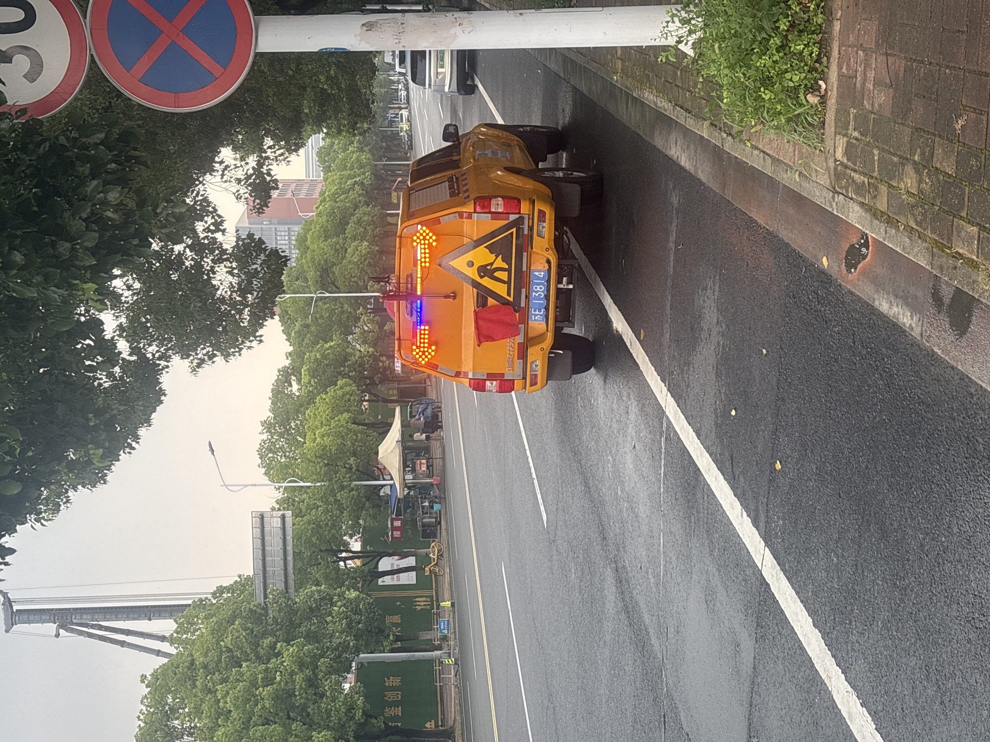 An orange utility vehicle, equipped with flashing amber and blue lights, is parked on the right side of a wet asphalt road. The vehicle displays a triangular warning sign with a silhouette of a worker digging, and a license plate reading "苏E 13814". The road has a white lane marking. In the background, trees line the street, and buildings are visible further away. To the right of the vehicle, a white pole supports two traffic signs: one indicating a speed limit of 30, and another a red circle with a blue background and a red diagonal cross, signifying no stopping. A pedestrian walkway made of brick is also present on the right. The scene appears to be in Suzhou Industrial Park, China, given the presence of Chinese characters on the vehicle and the style of the signage.