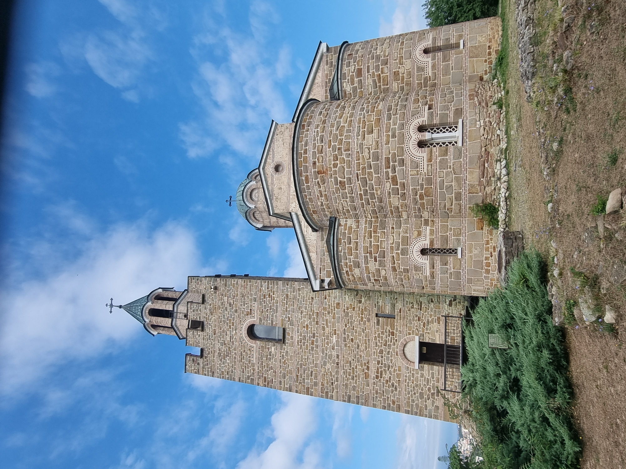The image shows the Tsarevets fortress in Veliko Tarnovo, Bulgaria, a medieval stronghold. The focal point is a stone church with a tall bell tower and a dome, characteristic of Orthodox Christian architecture. The building is constructed from large, rough-hewn stones, with decorative bands of smaller, lighter-colored stones. The bell tower has arched openings and a conical roof topped with a cross. The main church structure features arched windows and rounded apse-like sections. The sky above is a vibrant blue with scattered white clouds. In the foreground, there is dry, sparse vegetation and some scattered rocks, suggesting a hilltop location. A dense green shrub is positioned to the left of the church entrance. In the background, a distant cityscape can be seen, hinting at the broader location within the city of Veliko Tarnovo. There are no people or animals visible in the frame. The overall impression is one of historical significance and architectural grandeur against a clear sky.