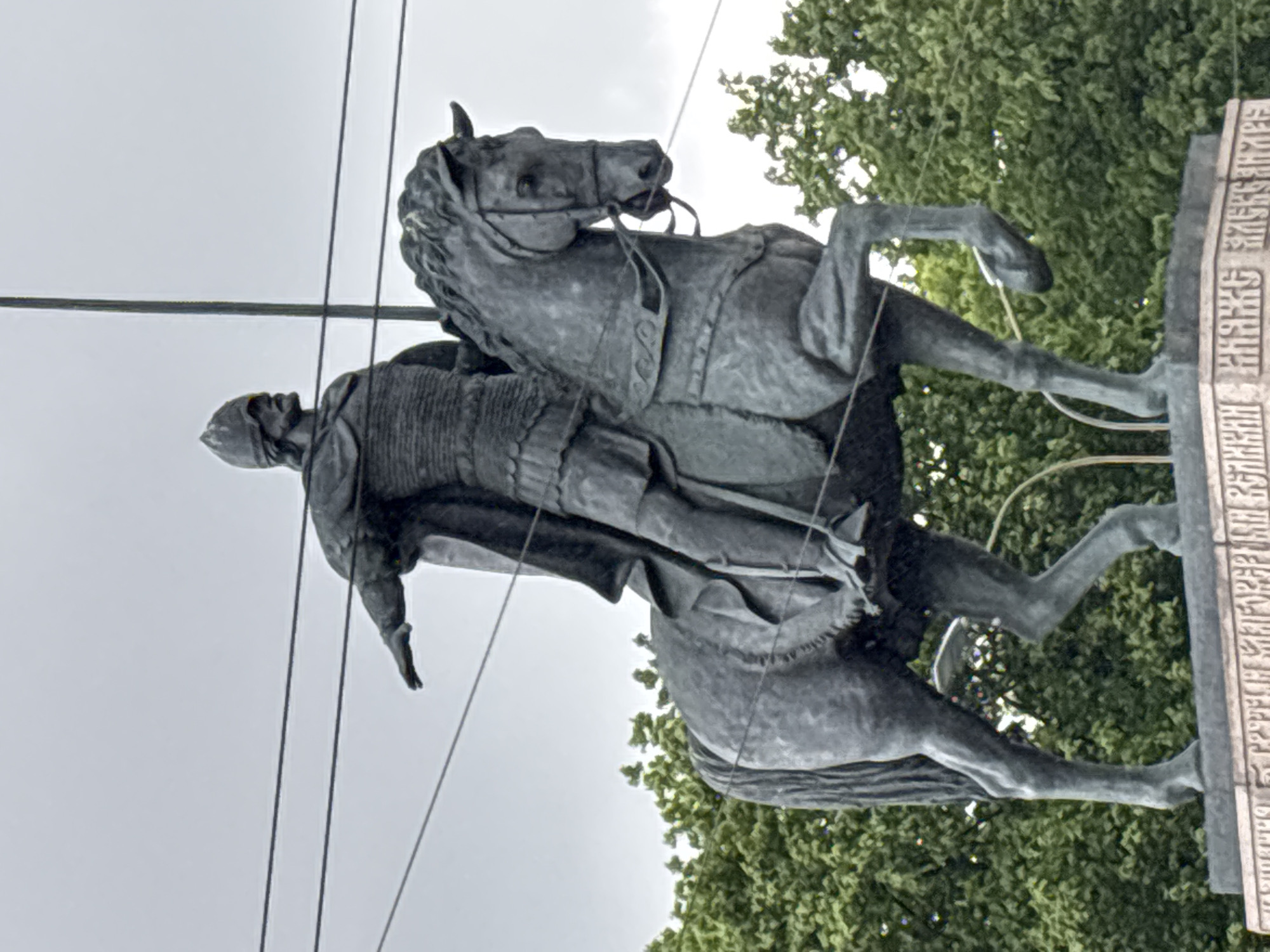 A statue of a mounted warrior is depicted from a low angle in front of a backdrop of leafy green trees and an overcast sky. The statue is constructed from a dark material that resembles bronze. The warrior figure is armored and helmeted and is mounted on a rearing horse. The statue is mounted on a plinth with inscriptions in Russian. Powerlines can be seen in the sky. This is the monument to Alexander Nevsky located in Saint Petersburg, Russia.