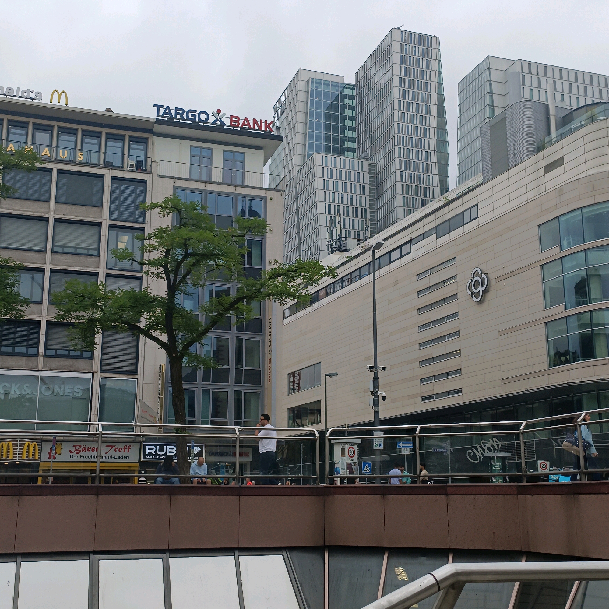 The image captures a street-level view of the banking district in Frankfurt, Germany, under an overcast sky. Dominating the background are several modern, high-rise office buildings with distinctive architectural styles, featuring extensive glass facades and a geometric arrangement of windows. One prominent building displays the "TARGOBANK" signage. In the mid-ground, a more historical building with a beige facade is visible, featuring large windows and balconies. A lush green tree with a substantial trunk branches out across the scene, partially obscuring the building facades.
At street level, a raised walkway with a metal railing separates the foreground from the mid-ground. Several individuals are present on this walkway. On the left, a man in a white shirt and dark pants stands with his hand to his ear, appearing to be on a phone call. To his right, two individuals are seated on a bench. Further to the right, a group of people are gathered, with one person walking away from the camera and another seated. Signs for businesses like "McDonald's," "Bären-Treff," and "RUB" are visible on the lower level behind the walkway.
Security cameras are mounted on a lamppost. The overall environment suggests an urban commercial area with a mix of contemporary and older architecture.