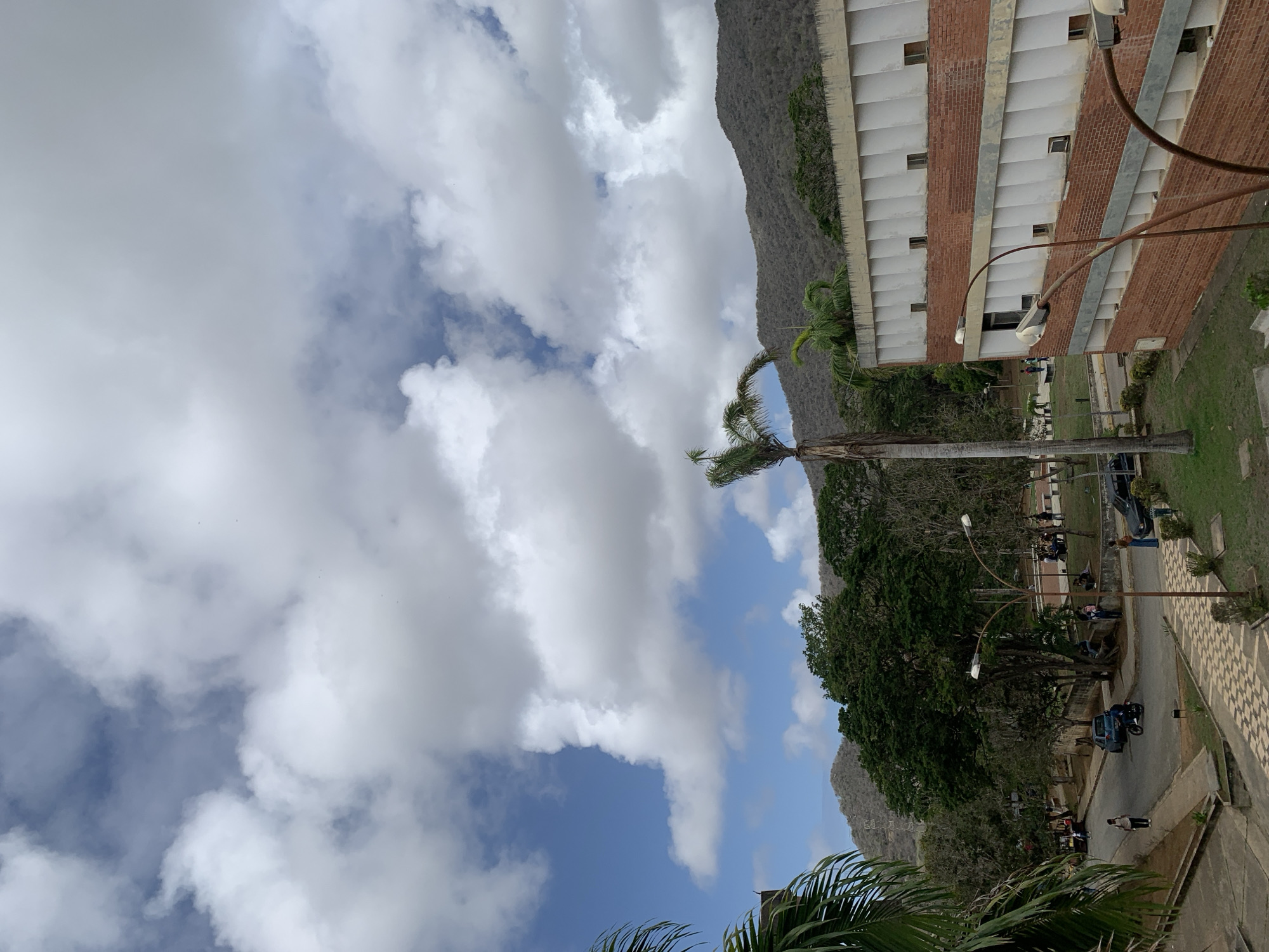 This media file depicts an outdoor scene in Las Piedras, Venezuela. A multi-story building with a brick and white facade dominates the right side of the image. In front of the building, a paved walkway with a checkered pattern runs along a grassy area. Several streetlights with curved poles are visible.
A road runs parallel to the walkway, with several parked vehicles, including a blue car and a motorcycle. People are seen walking and gathered in small groups on the road and the walkway, suggesting a normal day of activity. Lush green trees and a tall palm tree with a bare trunk are scattered throughout the scene, some providing shade. In the background, a rugged, gray mountain range rises against a sky filled with dramatic white clouds and patches of blue.
The lighting suggests it is daytime, likely late morning or early afternoon, given the brightness and the shadows. The weather appears partly cloudy, with the sun intermittently breaking through. The overall atmosphere is one of a relaxed, casual setting, possibly a campus or a public area. No visible text is present in the image.