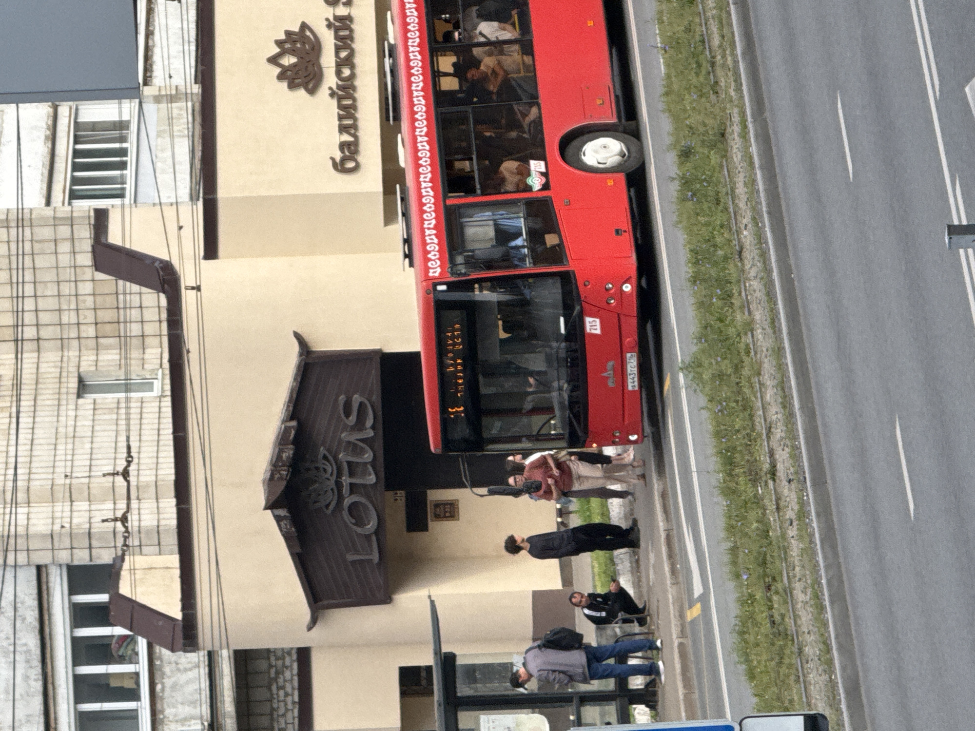 This image shows a red bus stopped at a bus stop in front of buildings with the signs "LOTUS" and "Балийский SPA" in Kazan, Russia. Several people are waiting at the bus stop, and others are visible inside the bus. The bus has the number "715" on its front and a license plate that reads "A443TC 716". The environment appears to be an urban setting with a street, sidewalk, and grass.