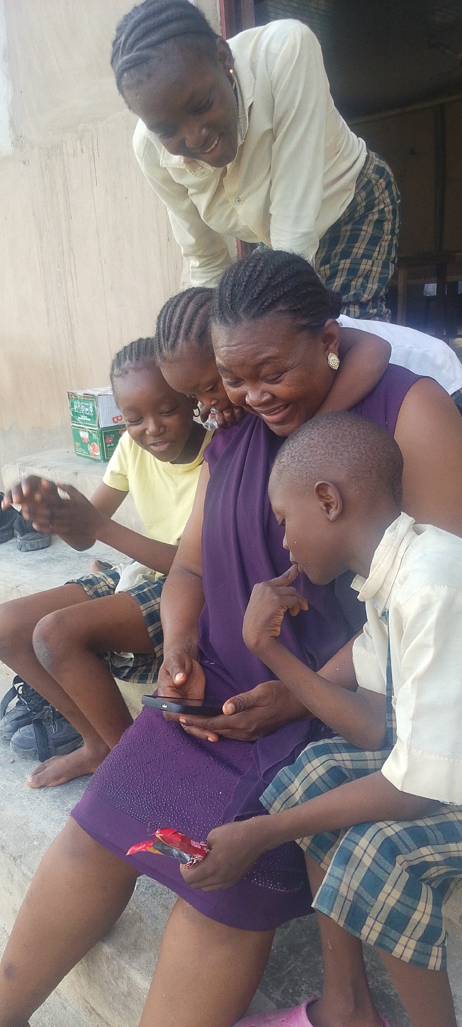A woman and four children are looking at a cell phone while sitting on steps in an outdoor setting in Lokoja, Nigeria. The woman, wearing a purple dress, is sitting with three of the children close to her. One child is on her lap. The children, a mix of boys and girls, are wearing casual attire and school uniforms. One child is holding a wrapped candy. Another woman is standing behind them, looking down and smiling. A green box of juice or drinks is visible in the background.