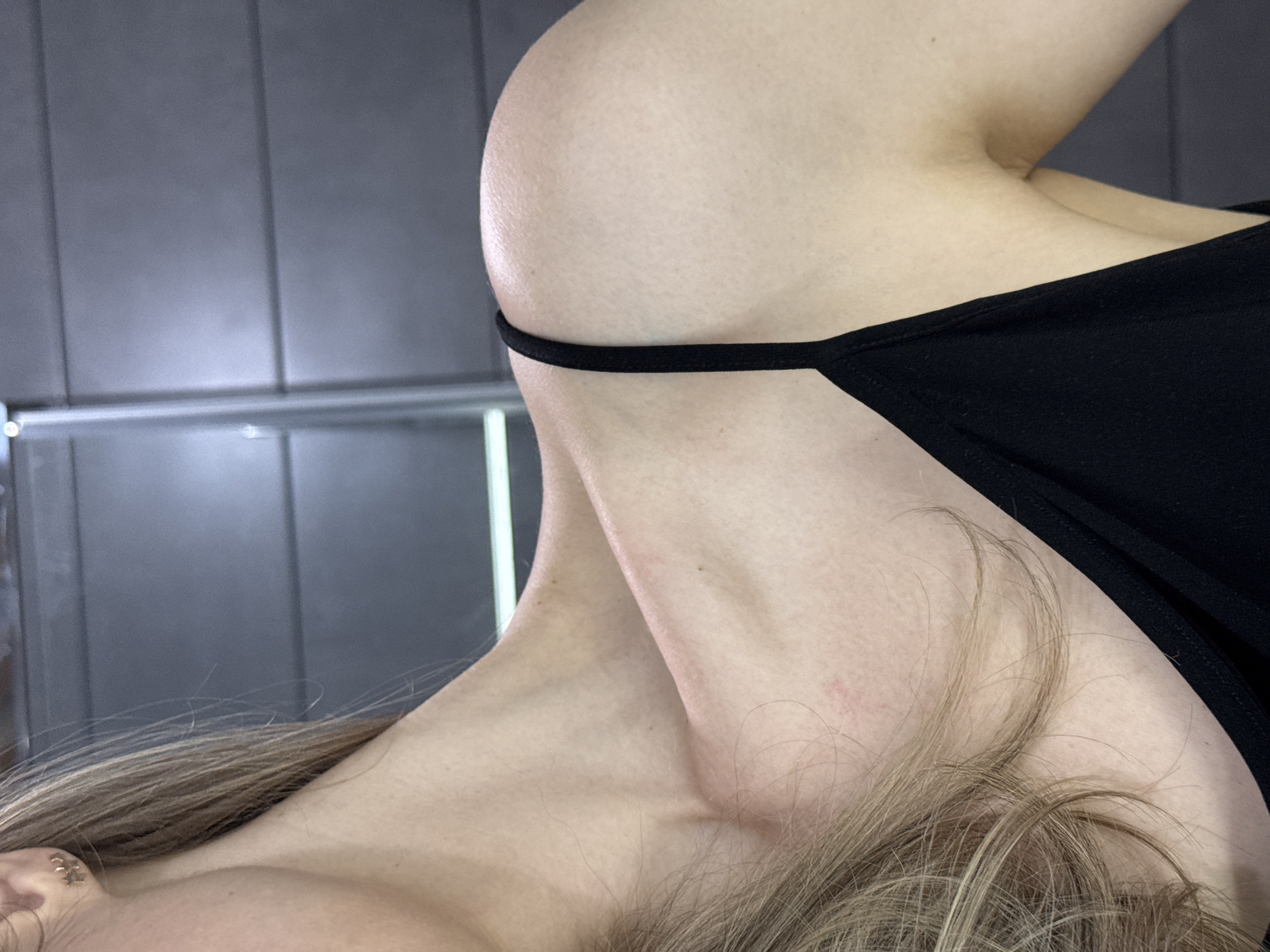 Close-up angled shot of a fair-skinned person. The focus is on their neck, shoulder, and part of their chest, partially covered by a black garment with a thin strap visible. Blonde hair falls in the foreground. The background is a blurred grey wall or paneling with a possible reflection. The location is not discernible from this image alone.