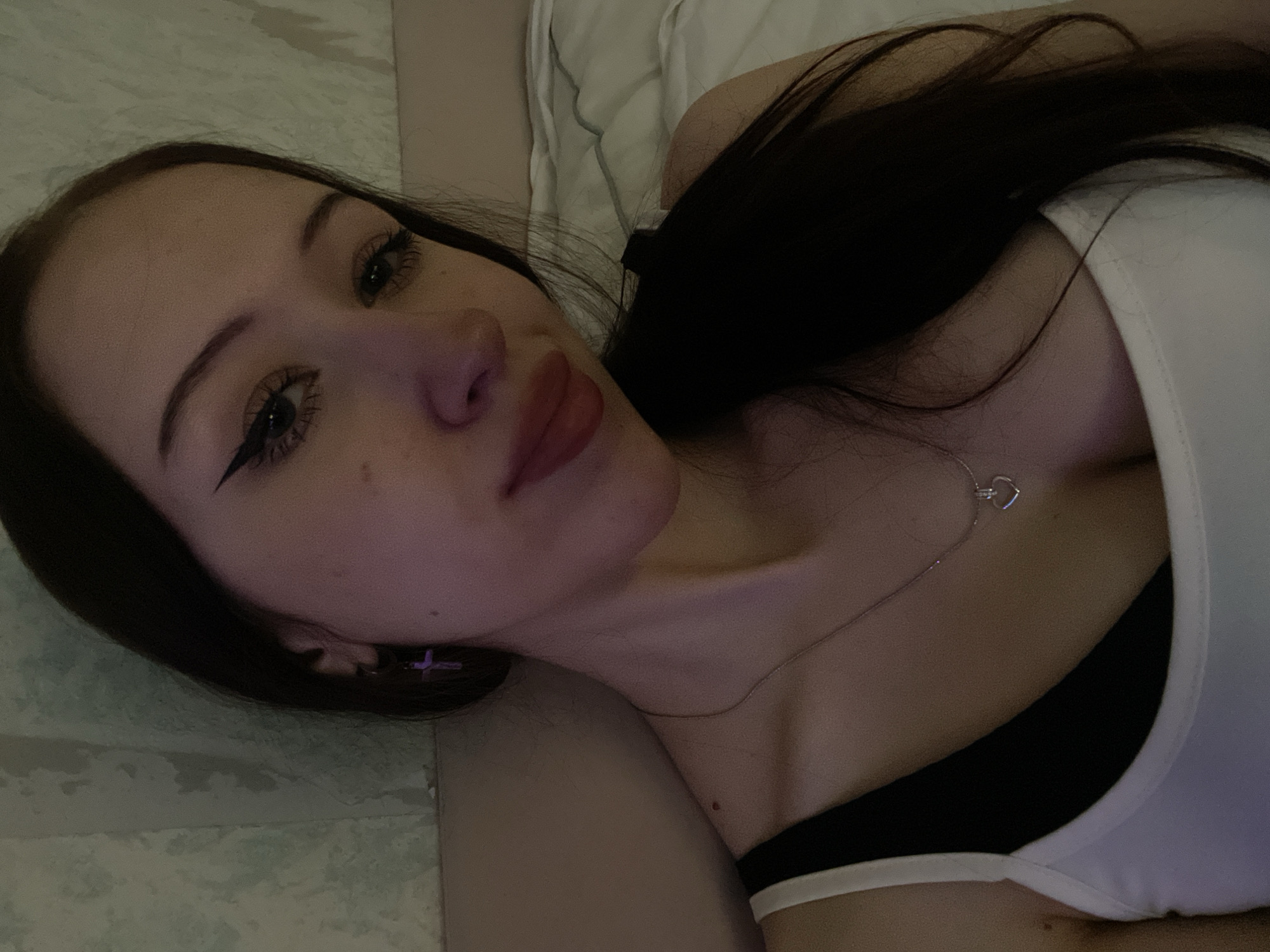 A young woman with dark hair, wearing a white tank top over a black bra, a silver heart necklace, and a purple cross earring, is shown in a close-up selfie. Her makeup includes winged eyeliner and lip gloss. The background appears to be a wall with textured wallpaper and a white duvet. The lighting is dim and warm, casting pink and yellow tones on her skin. The location is stated to be Votkinsk, Russia.