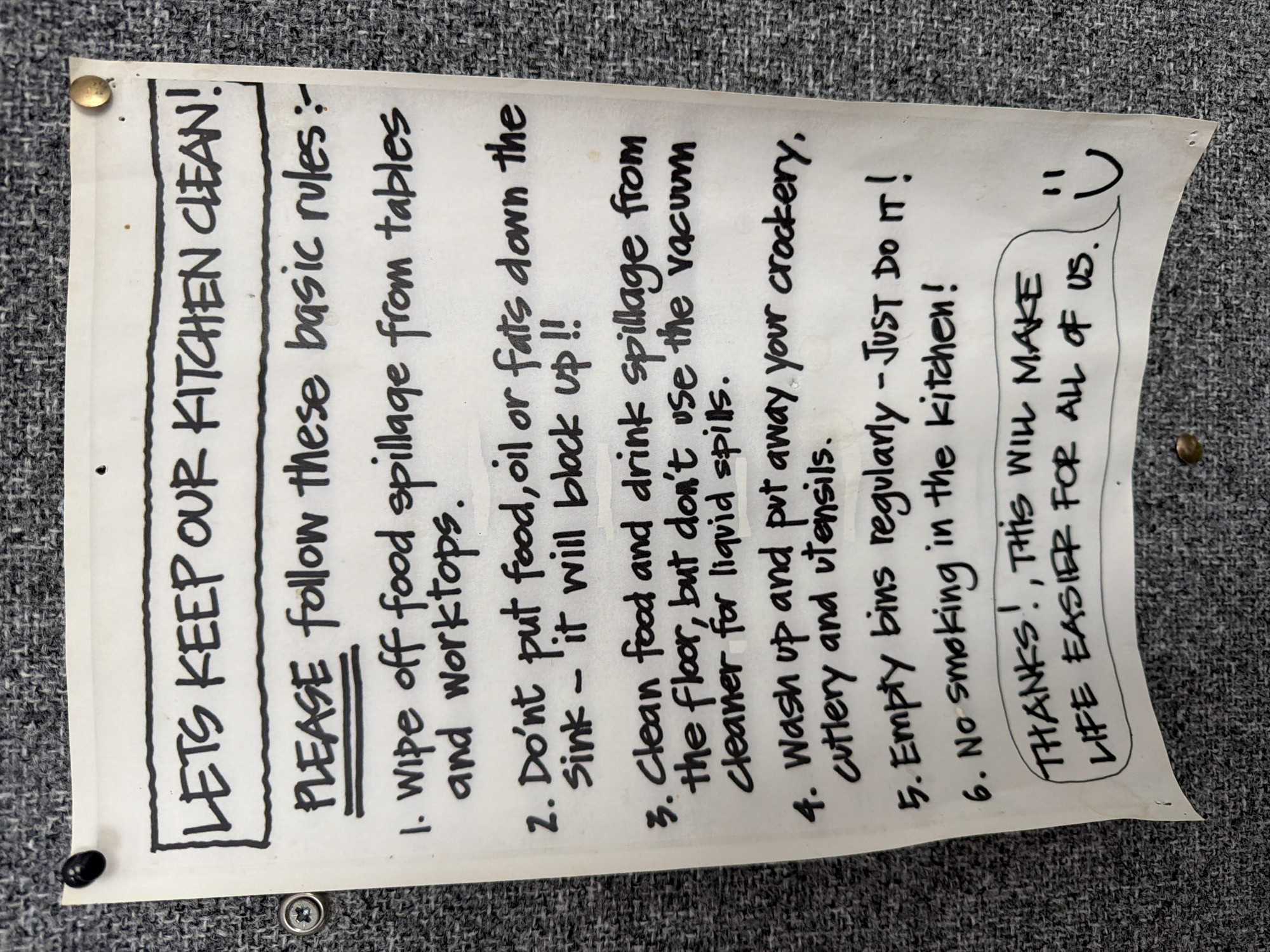 A handwritten note is pinned to a textured grey wall. The note, written in black marker, lists six rules for keeping a kitchen clean. The title, in large block letters, reads "LETS KEEP OUR KITCHEN CLEAN!". Below this, in smaller but still prominent letters, is "PLEASE follow these basic rules:". The rules are numbered and begin with "1. Wipe off food spillage from tables and worktops." and continue with instructions to avoid putting food, oil, or fats down the sink, to clean spills from the floor without using a vacuum cleaner, to wash up and put away crockery and utensils, to empty bins regularly, and a prohibition against smoking in the kitchen. The note concludes with a circled message that reads "THANKS! THIS WILL MAKE LIFE EASIER FOR ALL OF US." and a smiley face. The note appears to be in a communal area, possibly an office or staff kitchen. There is no information in the image that places it specifically in Epsom and Ewell, United Kingdom.