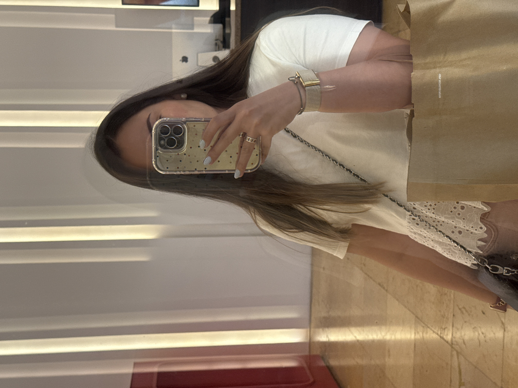 A woman is taking a selfie in a reflective surface. She is wearing a white shirt and a watch. She is holding a phone with a clear case and a brown paper shopping bag is visible. The location appears to be a store or shopping mall, judging by the internal architectural details. The exact location in Puebla, Mexico is not discernible from the image.