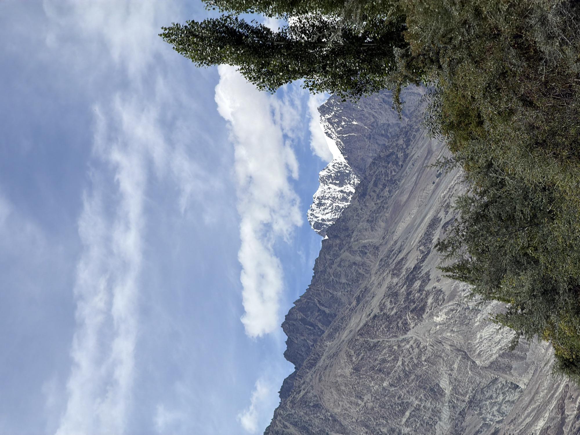 The image depicts a mountainous landscape with a clear sky. In the foreground, there are lush green trees and shrubs. Behind them, rugged, rocky mountains rise sharply. Some of the higher peaks are covered in snow, indicating a high altitude. The sky is a bright blue with scattered white clouds. The scene appears to be an outdoor natural environment. There are no people or man-made structures visible in the image. The location is identified as Terisha, India.