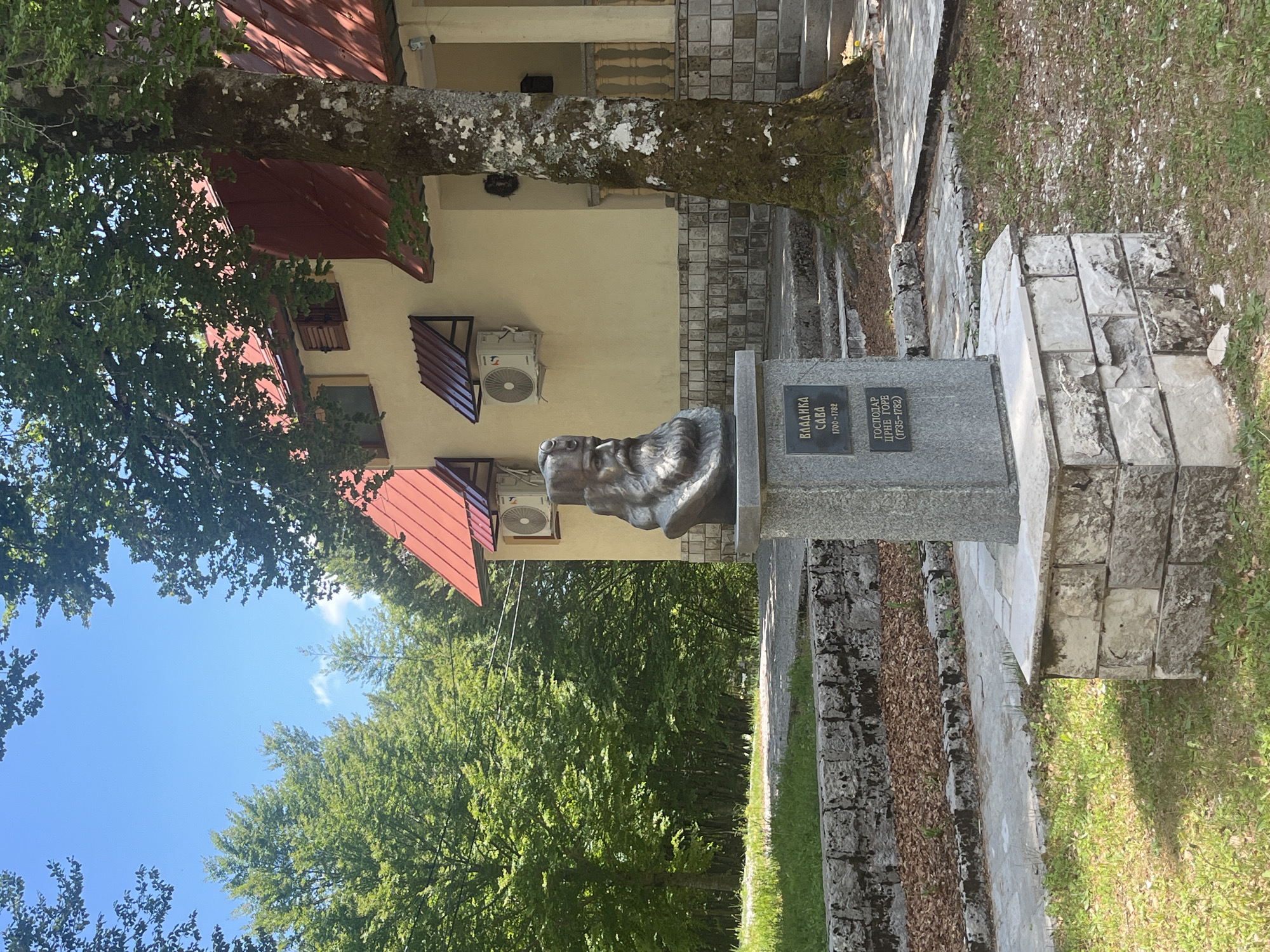 This is a photograph of a bronze bust of a man on a stone pedestal in Velji Bostur, Montenegro. The bust is centered in the frame and depicts a man with a beard. Inscription plates on the pedestal say "Владика Сава 1700-1782" and "Господар Црне Горе (1735-1782)". Behind the bust is a two-story building with a red roof and cream-colored walls. An air conditioning unit is attached to the building. A tree is positioned between the bust and the building. The surrounding area includes greenery, grass, and stone pathways. The sky is visible in the upper left of the frame.