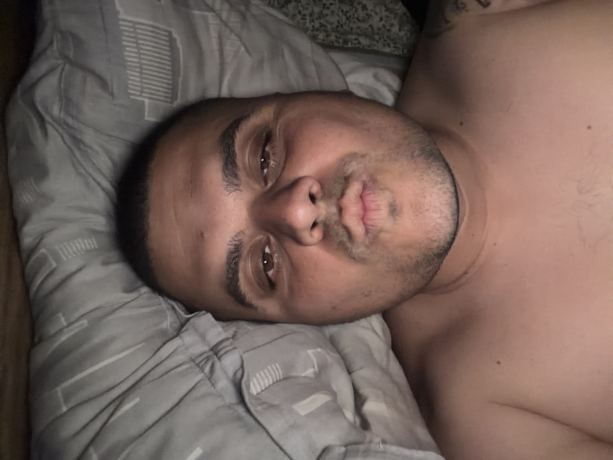 The image presents a close-up, horizontal portrait of an adult individual, likely male, lying down indoors. The person's head is resting on a light grey pillow adorned with a repeating geometric pattern of lines and rectangular shapes. The dim lighting suggests it might be evening or night, or a dimly lit room.
The subject has very short dark hair or a shaved head, and stubble or a short beard. They possess dark brown eyes, which are open and looking directly at the viewer. Their lips are distinctly pursed, forming a playful or pouting expression. The person's bare upper chest and shoulder are visible on the right side of the frame, showing light body hair and a few moles. A faint, dark tattoo is partially visible on the upper right shoulder area. Behind the subject's head, another darker, patterned fabric is visible, possibly part of the bed's sheets or duvet. There is no discernible text. The overall scene conveys a relaxed and intimate moment, with the subject engaging playfully with the camera.