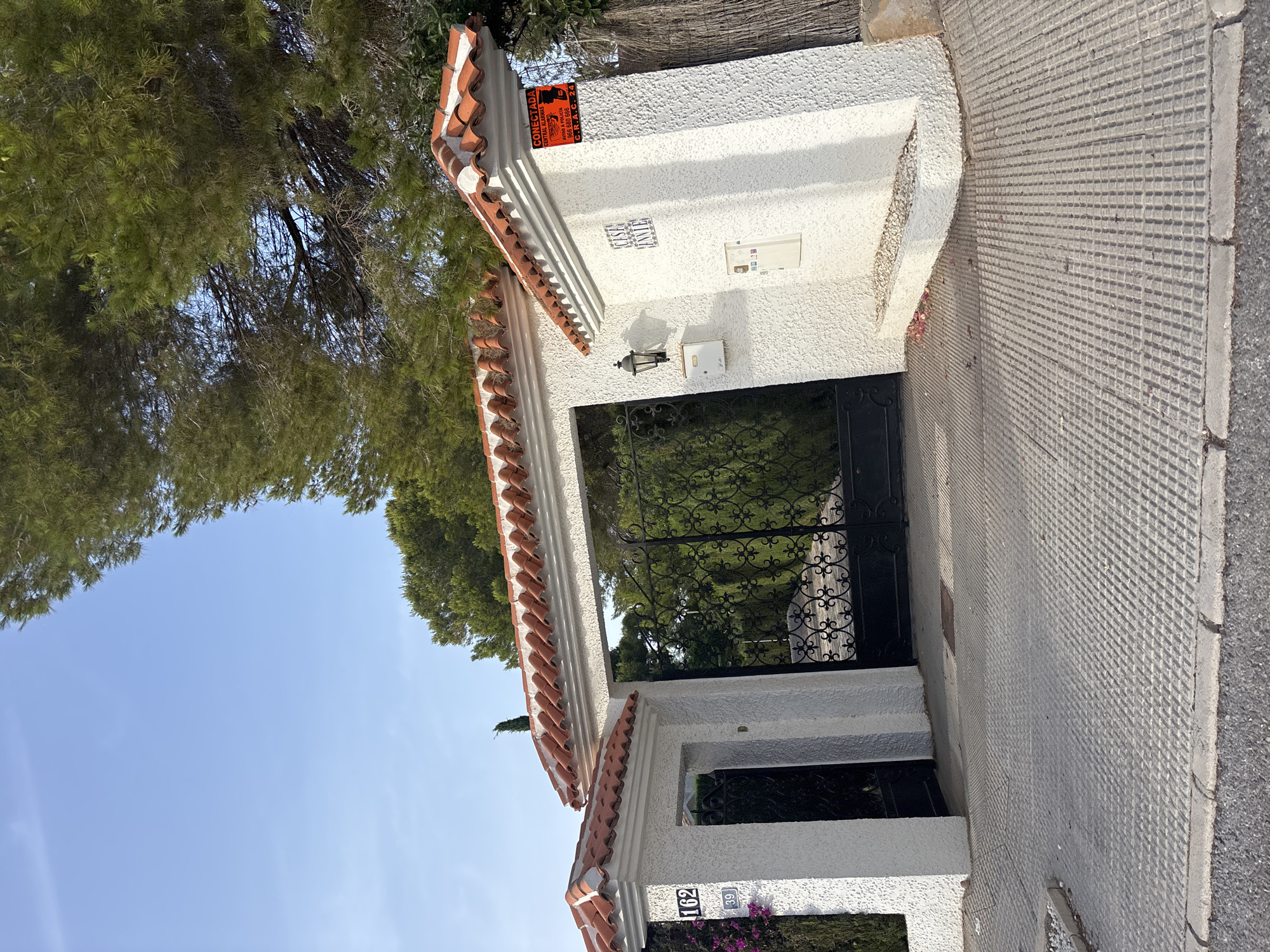 A security camera is mounted on a textured white wall to the right of an ornate black metal gate. The wall has a red-tiled roofline and a sign that reads "CONECTADA CENTRAL ALARMAS". The gate leads to a property with lush green foliage visible behind it. To the left of the gate, a white pillar displays house numbers "162" and "39". The ground in front of the gate is paved with textured concrete slabs. The sky above is clear and blue, with the top of a large pine tree visible on the right side of the frame. The scene appears to be an exterior view of a residential property's entrance in Orihuela, Spain.