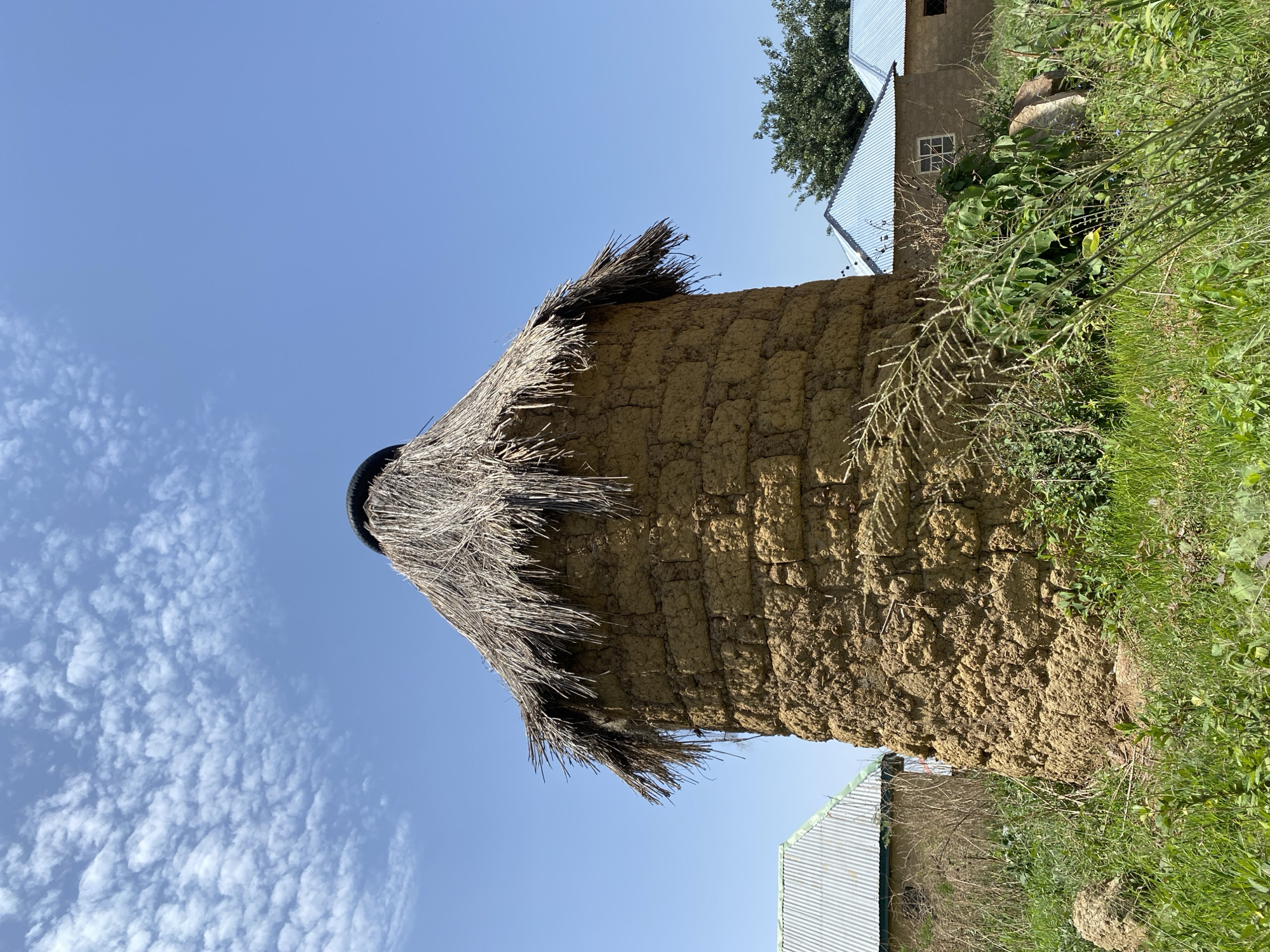 The image depicts a traditional mud granary structure with a thatched roof, a common feature in rural areas of Nigeria, including Durimi. The granary is constructed from mud bricks and topped with a thatched roof for protection from the elements. The structure is surrounded by vegetation, suggesting a rural setting. In the background, there are buildings with corrugated metal roofs, a sign of more modern construction. The sky is clear with some scattered clouds, indicating a sunny day.