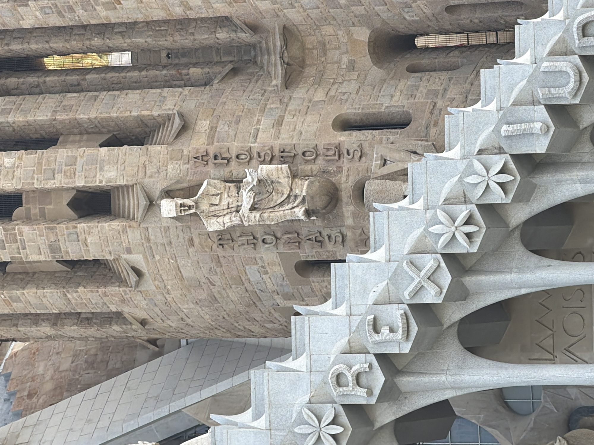 The image shows a close-up of the Sagrada Familia basilica in Barcelona, Spain. The architectural style is Gothic Revival, with intricate stone carvings. In the foreground, large, stylized letters are carved into the stone facade, forming the word "REX" and possibly parts of other words like "JESUS" and "LUX". These letters are part of a decorative staircase or ramp leading up the side of the basilica. Above and behind these letters, a stone sculpture of a seated figure, likely an apostle, is visible. To the right of the sculpture, the word "APOSTOLS" is carved vertically into the facade. The stone building is made of light-colored, rough-hewn blocks, with some areas showing smooth, polished stone. Sunlight illuminates the scene, casting shadows and highlighting the textures of the stone. The overall impression is one of monumental and detailed religious architecture.