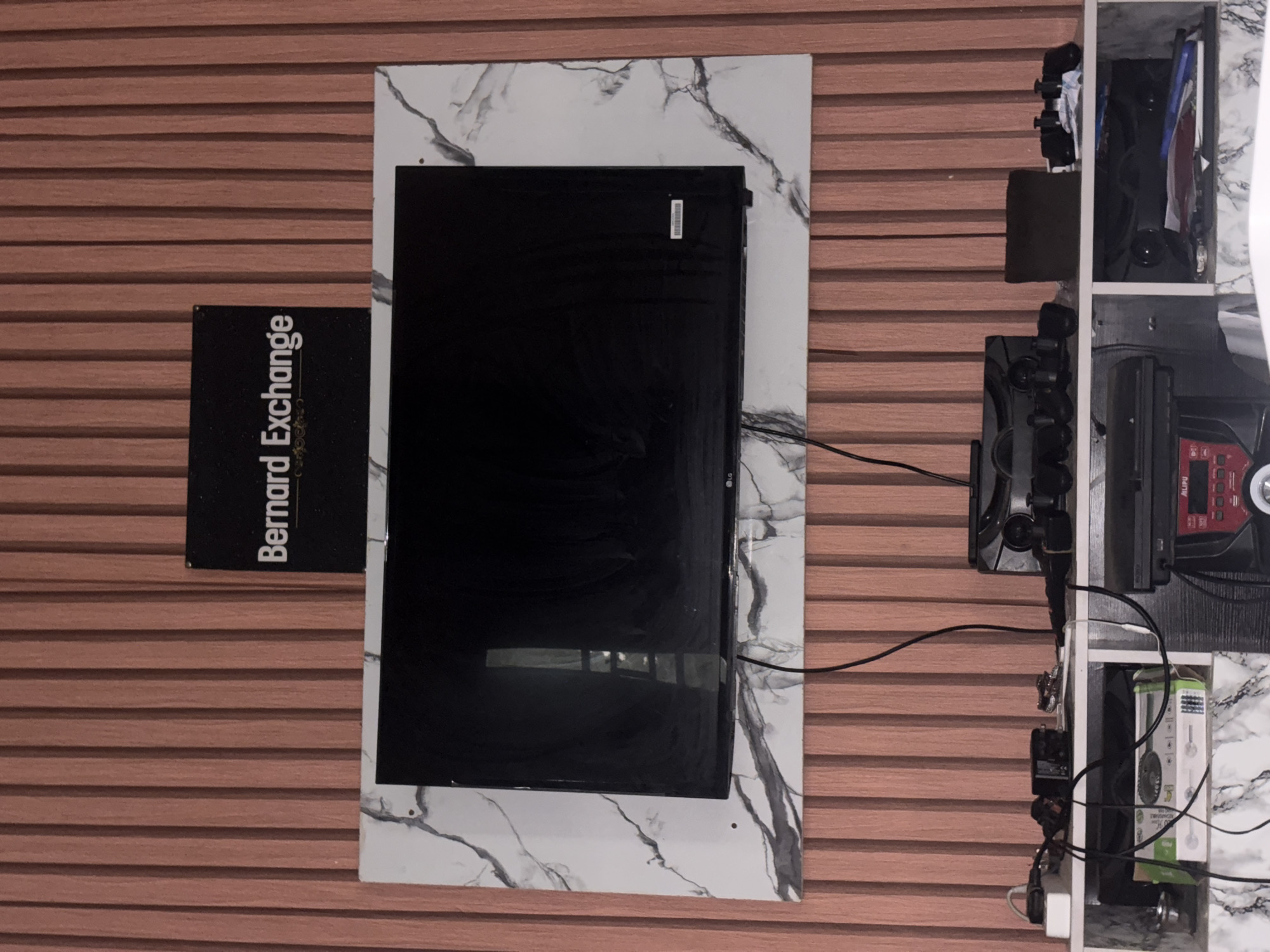 The image shows an indoor scene featuring a wall with horizontal wood-like paneling. On the wall is a flat-screen television mounted on a marble-patterned back panel. To the left of the TV is a black sign with white lettering that reads "Bernard Exchange" vertically. To the right of the TV, there is a white shelf unit filled with electronics and accessories, including speakers, a radio, and boxed items. Electrical cables are visible connecting the devices. The location is Ikot Ekpene, Nigeria.
