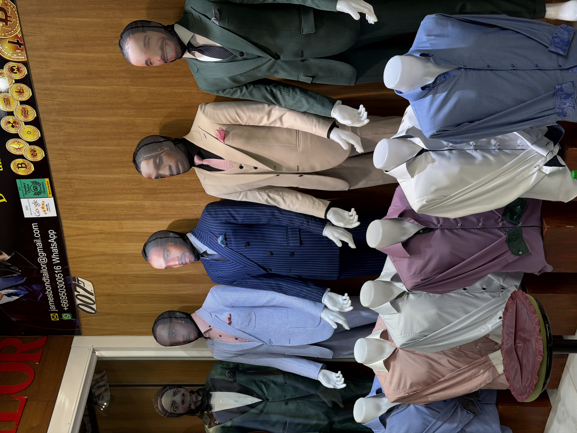 The image shows a tailor shop display in Chiang Mai City Municipality, Thailand. Five mannequins wearing suits and shirts are arranged in a row, each with a face-printed stocking over their heads. From top to bottom, the mannequins are dressed in a dark green suit, a beige suit, a pinstripe blue suit, a light blue suit, and a dark green suit with a white shirt and dark necktie. Next to the mannequins is a vertical sign with the business name "JAMES BOND TAILOR" alongside the email address "jamesbondtailor@gmail.com" and a WhatsApp number (+66950300516). The sign also shows the "007" logo. The sign features awards from TripAdvisor and Google, indicating "Travellers' Choice 2023" and "Best of the Best" respectively. The display background appears to be a light wooden wall.