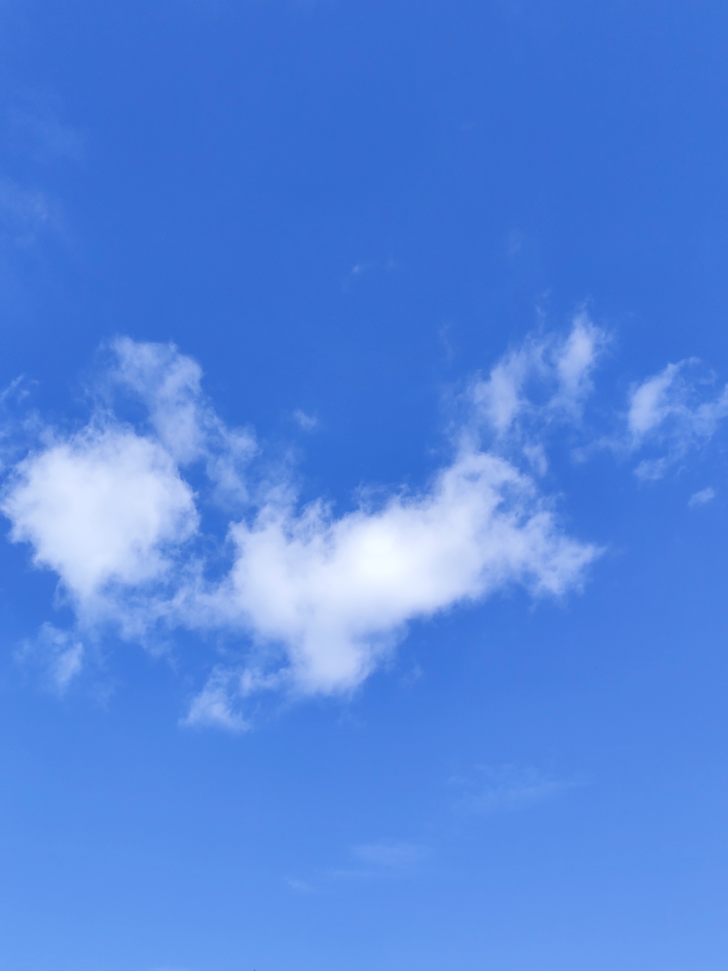 A bright blue sky is filled with scattered white clouds. A large, wispy cloud formation stretches horizontally across the mid-section of the frame, with smaller, more defined clouds to its left and right. The image appears to be a daytime shot taken outdoors, with no specific landmarks or identifiable structures from the city of Барлакский, Russia visible.