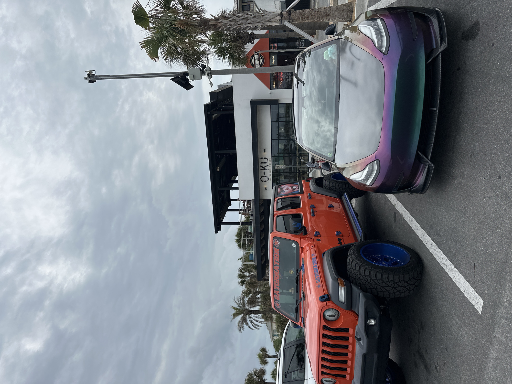 The image shows two cars parked outside the restaurant "O-Ku" in Jacksonville Beach, Florida, United States of America. The sky is overcast. There is a white car, an orange Jeep with a "GATORLATION" decal, and a color-shifting (possibly iridescent) Tesla Model 3. The restaurant has a black awning and glass doors, with outdoor seating visible on a patio. A palm tree is visible next to the building. A pole with a camera and light fixture is visible above the cars. "UKUMADES" is written on an orange sign next to the restaurant.