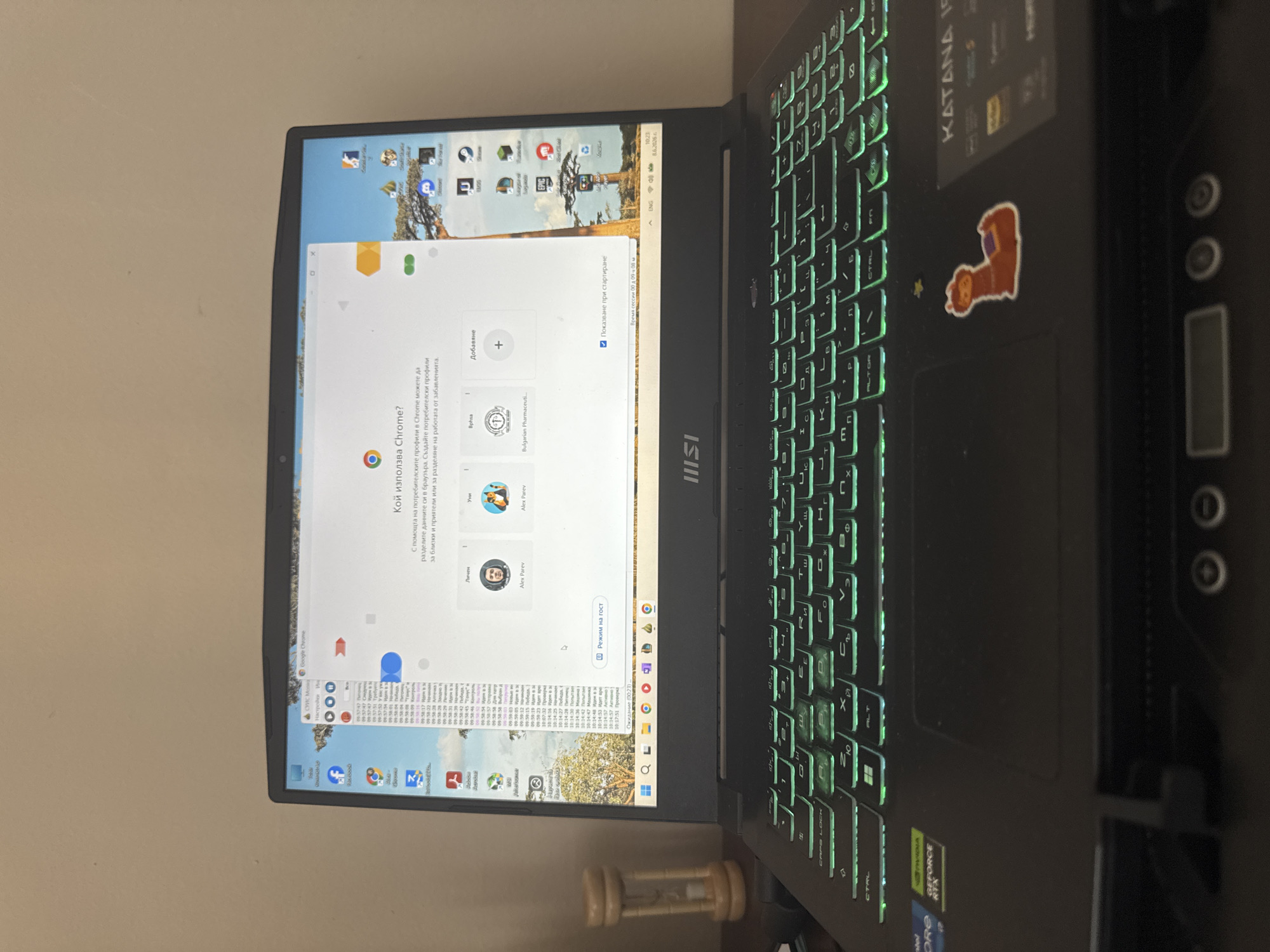 The image shows a laptop with a green-backlit keyboard, open and displaying a Google Chrome browser window. The browser window shows a prompt asking "Who is using Chrome?". Below this, there are options to add user profiles with avatars and names. The operating system appears to be Windows, with a taskbar at the bottom displaying various icons, including search, file explorer, and several application shortcuts. The desktop background shows a scenic view of a savanna with trees and a distant mountain. On the left side of the laptop screen, there is a vertical bar with icons for popular applications like Facebook and other social media platforms. A small hourglass timer sits on the desk to the left of the laptop. The overall environment suggests a personal workspace. The provided crops highlight specific elements of the laptop screen and keyboard, as well as a sticker of a llama on the laptop's surface. There are no specific visual cues in the image that would place it in Sofia, Bulgaria.