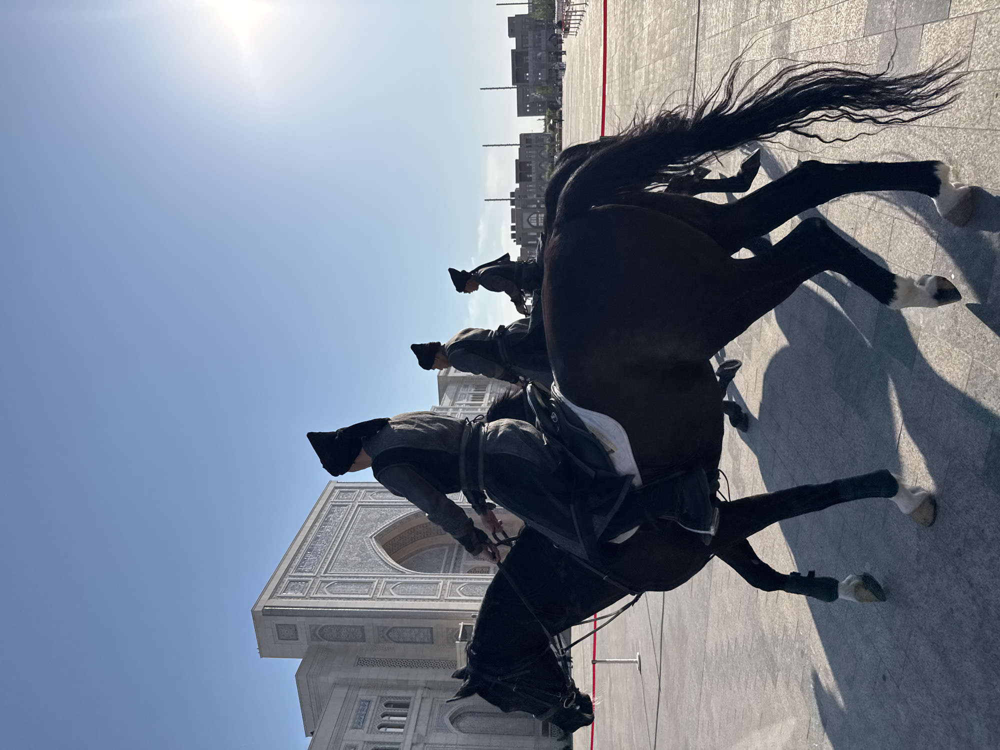 A medium shot captures a line of horse-mounted guards in Tashkent, Uzbekistan. The foremost guard, dressed in traditional garb and a tall black hat, sits atop a dark horse with white markings on its legs. The horse and rider are positioned to the left of the frame, with the rider facing slightly away from the viewer. Two more guards and horses are visible behind the first, diminishing in clarity with distance.
The guards and horses are situated on a paved plaza, likely a ceremonial ground. To the left of the guards, a large, ornately decorated white building with arched entrances and detailed tile work is prominent. The building's architecture suggests a historical or cultural significance. Behind the guards, in the distance, are modern buildings, possibly residential or commercial structures. The sky is clear and blue, indicating a sunny day. A faint red line is also visible on the paved ground near the horses.