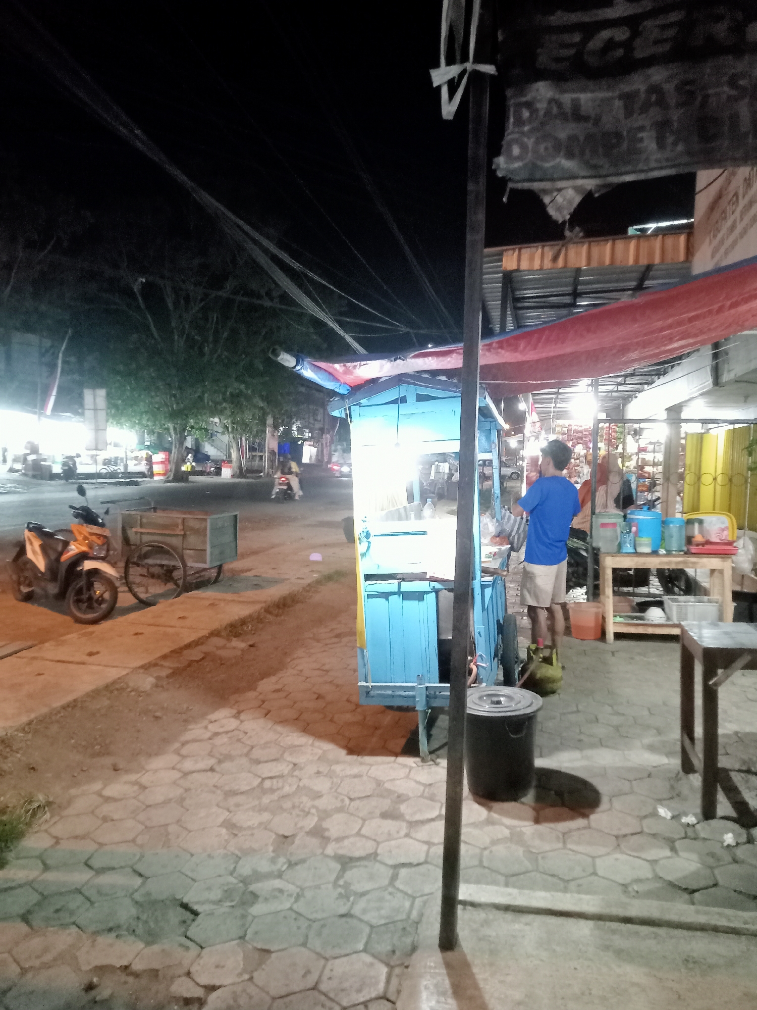 A nighttime street scene in Mangunsari, Indonesia features a blue food cart with a red and blue tarp overhead. A man wearing a blue t-shirt and khaki shorts stands behind the cart, appearing to prepare or serve food. To the left of the cart, an orange and white motorcycle is parked next to a wooden cart on wheels. The ground is paved with hexagonal tiles. In the background, trees line the street, and streetlights illuminate the area. Another food cart with a yellow tarp is visible further down the street, and several other carts and stalls are present. The overall impression is of a lively night market or street food area.