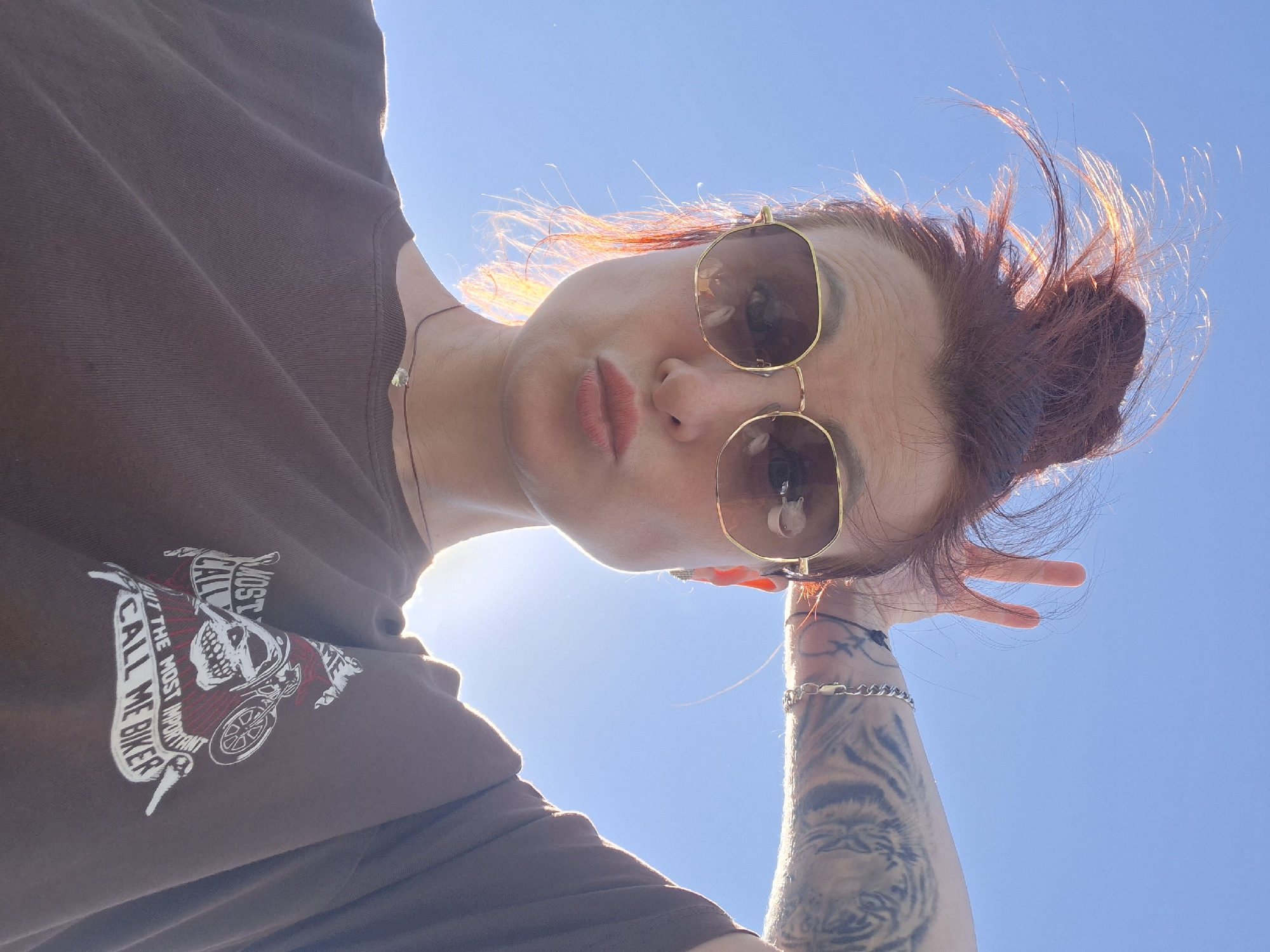 A woman with reddish-brown hair pulled into a messy bun, wearing sunglasses and a brown t-shirt, is captured in a low-angle, outdoor selfie. Her left hand is raised to her head, forming a bunny-ear gesture with two fingers. A tattoo of a tiger is visible on her left forearm, and she is wearing a silver bracelet. A delicate necklace with a small pendant is visible on her neck. The t-shirt has a graphic print featuring a skull and motorcycle with text that reads "MOST CALL ME BUT THE MOST IMPORTANT CALL ME BIKER." The background is a clear blue sky, illuminated by bright sunlight, suggesting a daytime setting. The provided location of Смалявічы, Belarus, is noted.