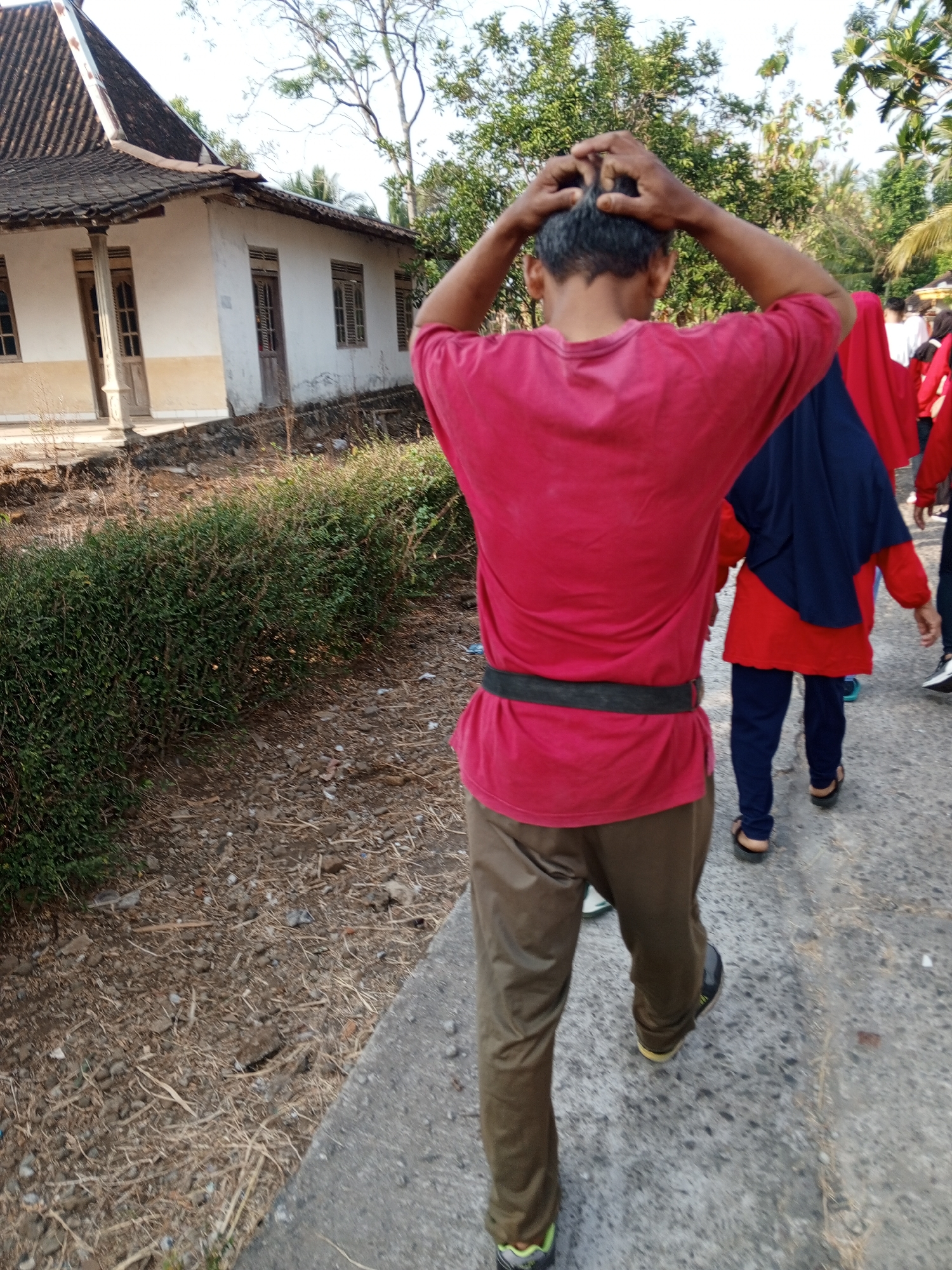 In Lemahbang, Indonesia, an individual wearing a red t-shirt and brown pants is walking on a paved path, their back to the camera. They are holding their hands to the back of their head, as if adjusting their hair. To the left, a white building with a dark tiled roof and wooden window frames is visible. A dense green hedge runs along the side of the path, separating it from the building. Further down the path, other individuals, some wearing red and blue head coverings, are also walking. The ground is covered with dry leaves and small rocks. The scene appears to be outdoors, with trees and foliage in the background under a clear sky.
