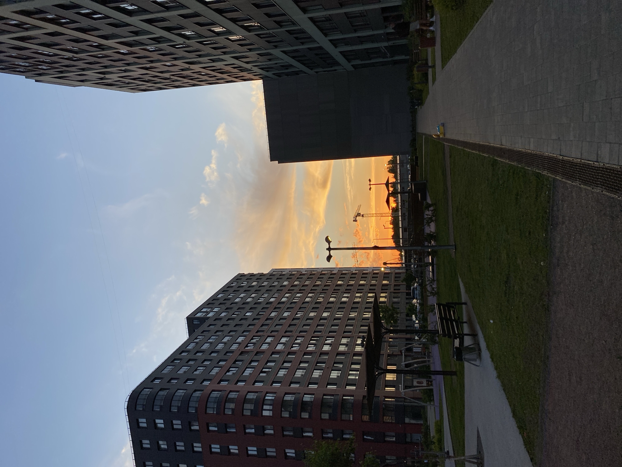This image depicts a modern urban landscape at sunset in Saint Petersburg, Russia. The scene is framed by tall, contemporary buildings and features a walkway and green space.
In the foreground, a paved walkway made of rectangular concrete or stone tiles stretches into the distance, bordered by a strip of manicured green grass. A long, dark metal grate separates the walkway from the grass. Further along the grass, two dark park benches are visible, suggesting a public amenity area.
On the left side of the frame, a multi-story modern building with a distinctive architectural style stands prominently. It features a curved facade with a mix of dark gray and reddish-brown panels, and numerous rectangular windows. On the right, another large, dark building presents a more angular, shadowed face to the viewer.
Between these buildings, the sky is painted with the warm, vibrant hues of a sunset, transitioning from bright oranges, pinks, and yellows near the horizon to a softer blue higher up. Silhouetted against this colorful sky are several construction cranes, indicating ongoing development in the area. Multiple streetlights or park lamps are also visible, some appearing to be illuminated. Above, a third, darker building is partially seen at the top of the frame, showcasing the grid pattern of its windows against the lighter blue sky.
The weather appears clear with some scattered clouds catching the evening light. The overall mood of the image is serene and peaceful, capturing the quiet beauty of a city transitioning from day to night. There is no visible text in the image.