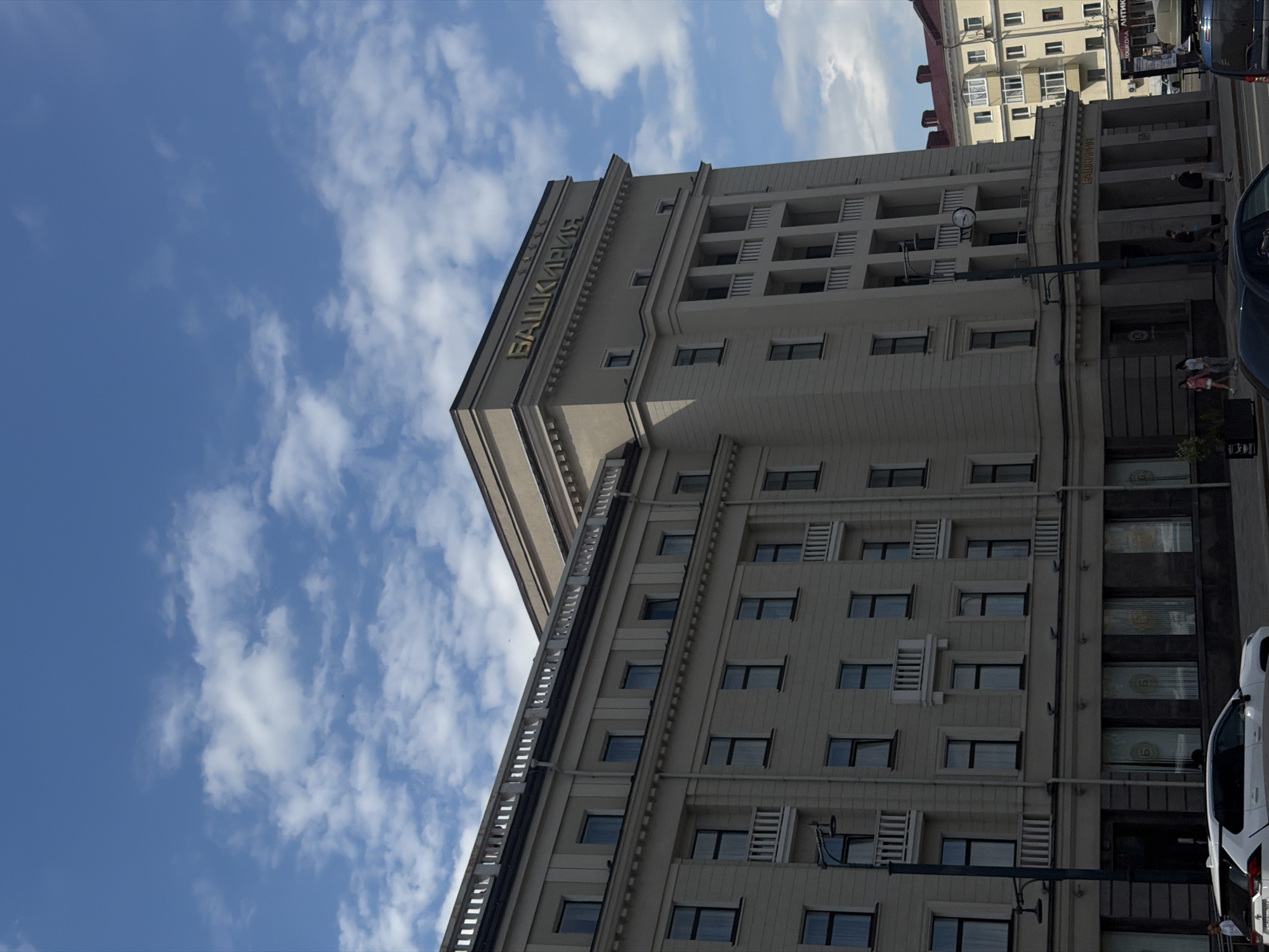 The image shows a low-angle shot of a modern, light-colored building with a blue sky and white clouds in the background. The building has multiple stories with many windows and balconies. Gold lettering is visible on the facade, reading "БАШКИРИЯ". A street is visible at the bottom of the frame with a few people and vehicles, including a white van. The scene appears to be set in a city, possibly during the daytime, with mild weather indicated by the presence of light clouds.