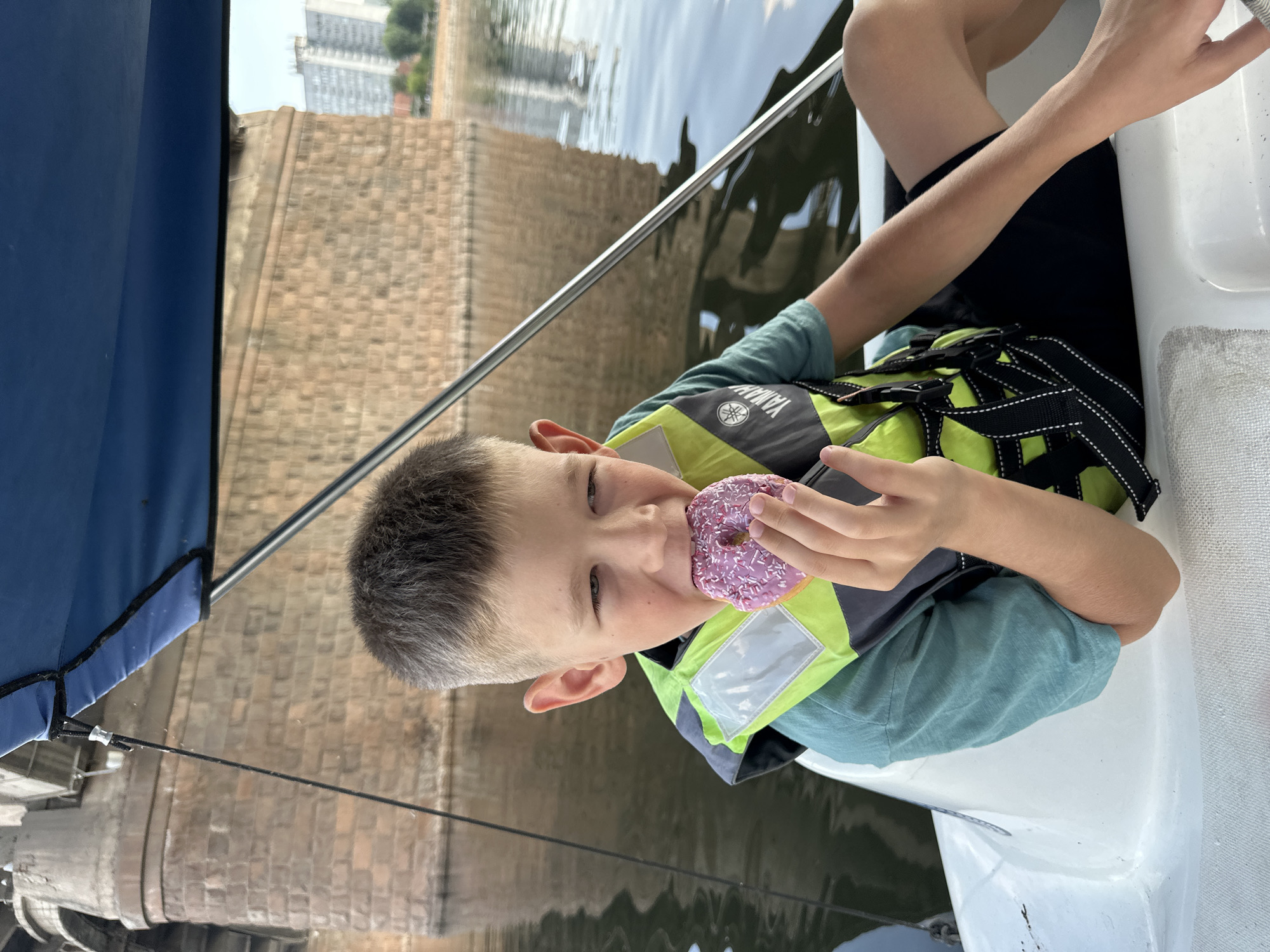 A young boy wearing a teal t-shirt and a neon yellow and black life vest is sitting on the edge of a white boat. He is holding a pink frosted donut with purple sprinkles to his mouth and appears to be taking a bite. The boy has short, greyish-brown hair and is looking to his right, with his left eye partially closed. His right hand is holding the donut, and his left arm is bent. The boat is on a body of water, with a stone bridge and buildings visible in the background. The water is dark and reflective, and there is a blue canopy overhead. The overall scene suggests a leisurely activity on the water.