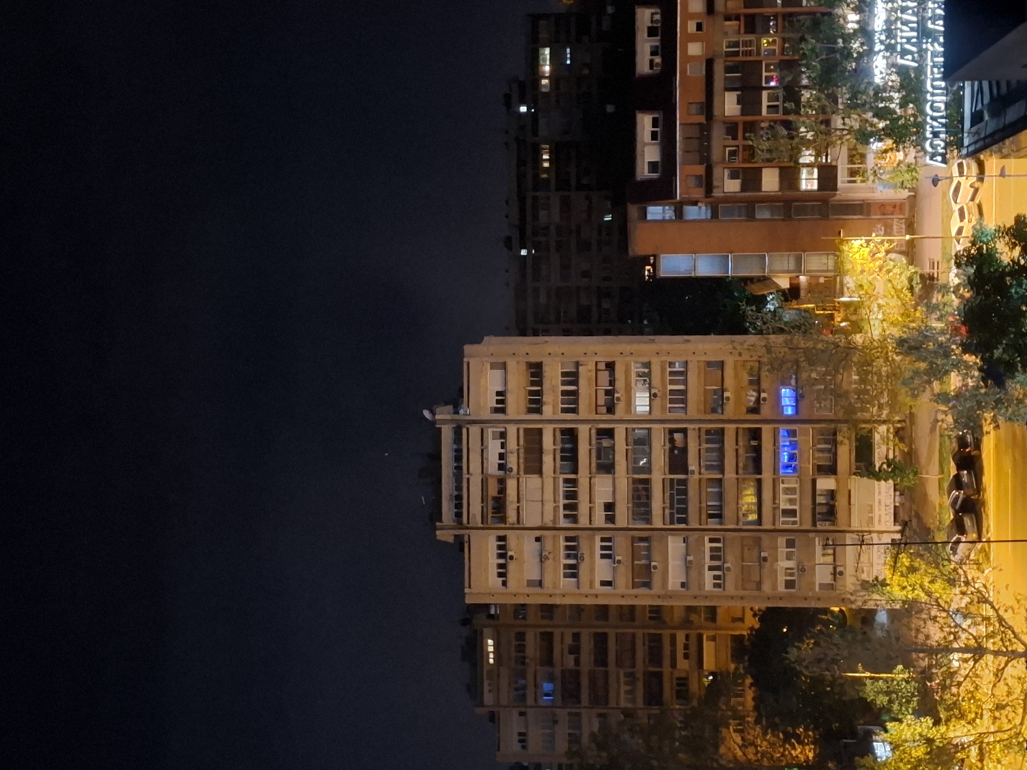 The image shows a night scene in Novi Sad, Serbia. A multi-story residential building with numerous windows is illuminated by streetlights. To the right are other buildings and the illuminated signage of "ATD Vojvoda Stepa". There are trees and streetlights providing a golden glow to the scene. People appear to be walking or standing near parked scooters on the sidewalk. To the left, the scene fades into the darkness of the night sky. The building in the foreground is set against the backdrop of a dark sky and illuminated city lights.