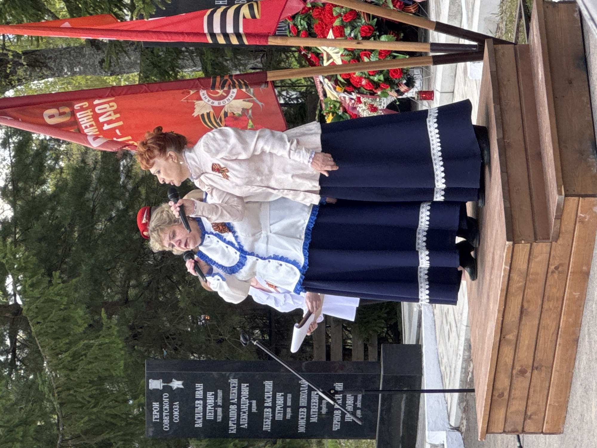 Two women stand on a wooden platform, each holding a microphone and speaking. They are wearing matching outfits of white blouses with decorative trim and long navy skirts with white trim. Behind them are red flags with Soviet-era emblems and black and gold stripes. A flower arrangement is visible near the flags. In front of the platform is a black memorial stone with inscriptions in Cyrillic, listing the names of heroes of the Soviet Union. A green, wooded area is behind the memorial. This scene is located in Chuprino, Russia.