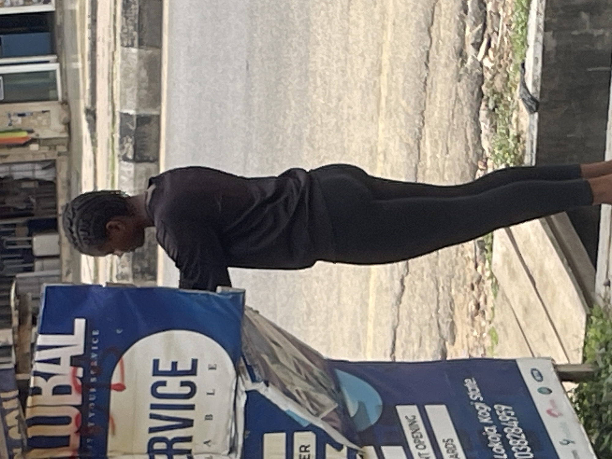 An outdoor medium shot shows a young woman with dark hair pulled back and braided, facing right, partially obscured by a large blue sign. She is wearing a black long-sleeved shirt and black leggings. The sign has white lettering and a circular graphic. The background reveals a street with a concrete curb and pavement, suggesting an urban setting. The lighting indicates it is daytime.