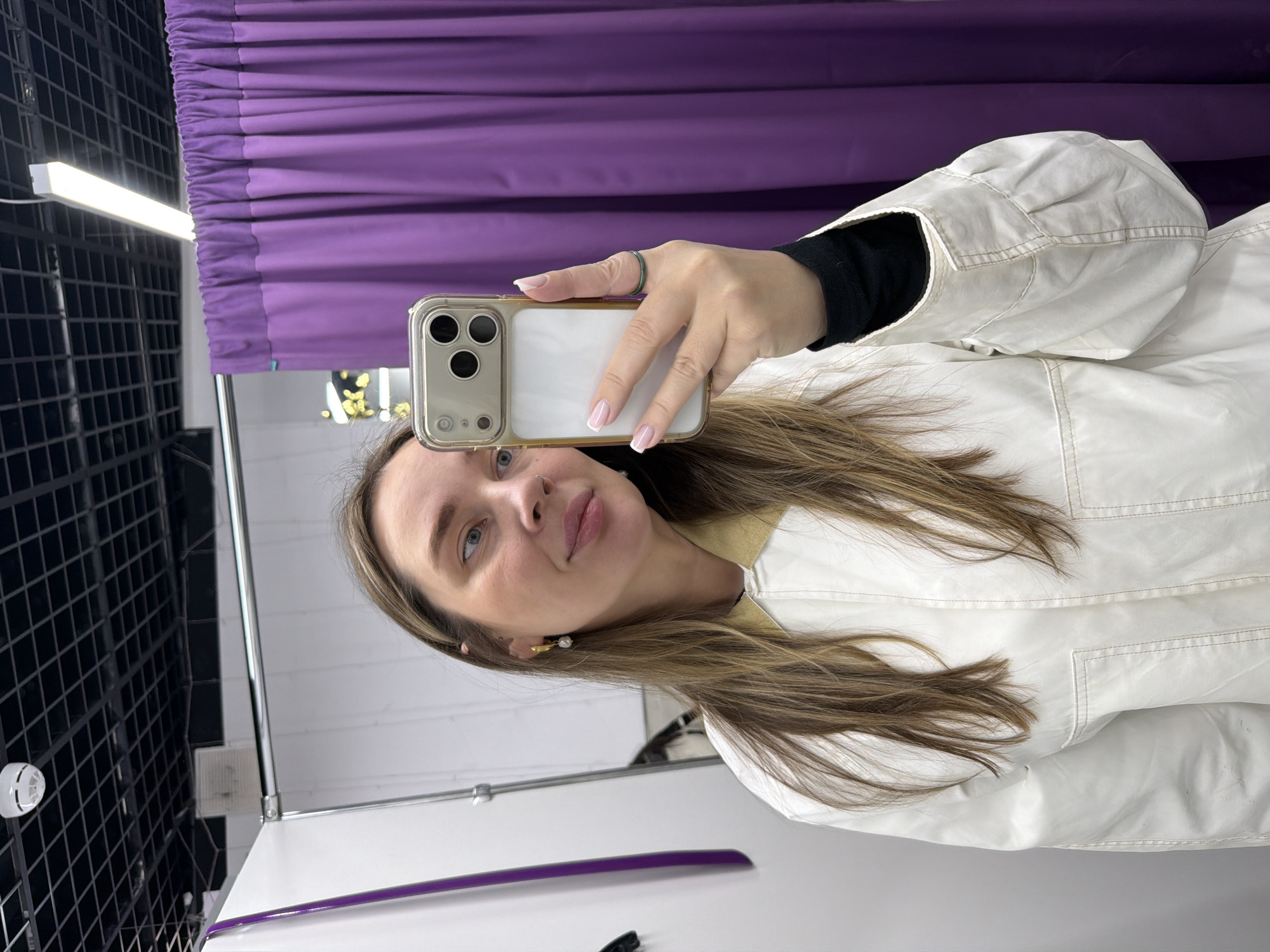 A woman is taking a selfie in what appears to be a dressing room. She is holding a smartphone with a visible camera array. The background includes a purple curtain and a black metal grid structure, possibly part of the dressing room's framework. The woman is wearing a white jacket and has long brown hair. There is a light fixture visible on the ceiling. There is no information to deduce if the photo was taken in the city of Moscow, Russia.