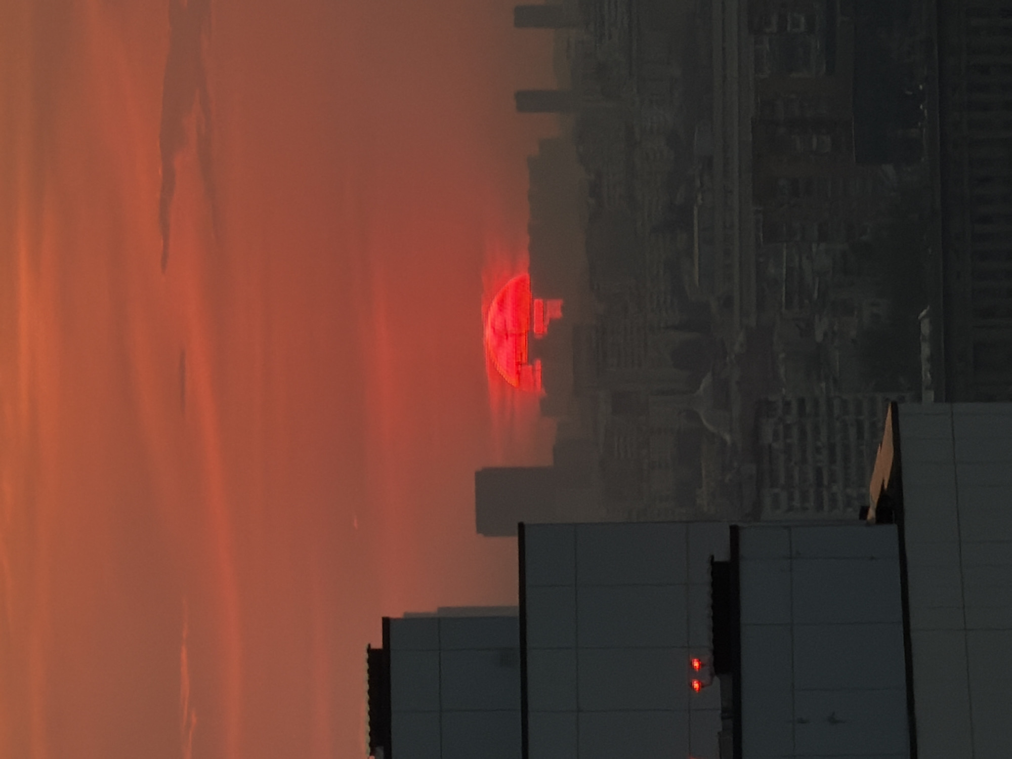 Here is a detailed description of the image:
**Environment:** The image captures an urban setting during what appears to be a heavily polluted or dust-filled sunset, casting an intense orange-red hue across the sky. The air is thick with particulate matter, obscuring visibility.
**Location:** The location appears to be an urban environment, likely a city, with a dense collection of buildings visible in the middle ground. No additional information available.
**Objects:**
* **Buildings:** Various types of buildings dominate the scene. In the foreground are the rooftops of tall, modern buildings with flat surfaces and geometric shapes. Further back, amidst the smog, are silhouettes of high-rise buildings, possibly skyscrapers, contributing to a dense cityscape.
* **Celestial body:** A partially obscured, intensely red sun (or a celestial body resembling a sun) hangs low in the sky. Its color is heightened by atmospheric conditions.
**Composition:** The photograph is composed with the foreground buildings providing a stark contrast against the polluted sky and the obscured cityscape behind. The unusual atmospheric conditions and the stark juxtaposition of the built environment create an unsettling atmosphere.