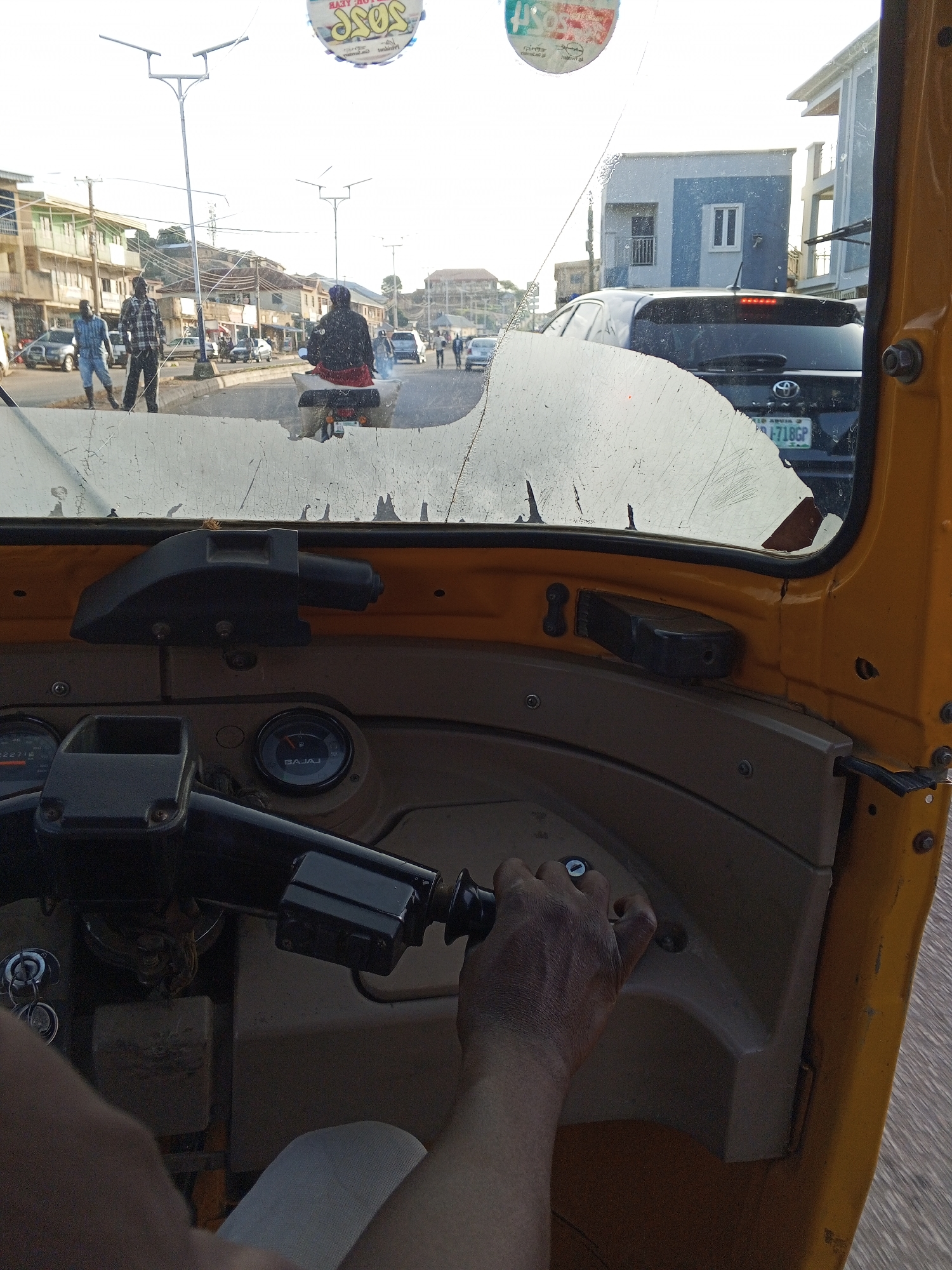 The image, captured from the interior of a yellow auto-rickshaw, depicts a street scene in Jos, Nigeria. The cracked windshield obscures a view of the road ahead, where traffic is present. On the right side of the windshield, a black SUV with a license plate reading "AID ABUJA F -718GP" is visible. In the mid-ground, two pedestrians are walking on the sidewalk, and several cars are on the road. Streetlights are visible along the road. The interior of the auto-rickshaw shows the steering wheel and a hand gripping it, indicating the driver's perspective. A fuel gauge labeled "BAJAJ" is also visible on the dashboard. The overall environment appears to be an urban setting with buildings lining the street.