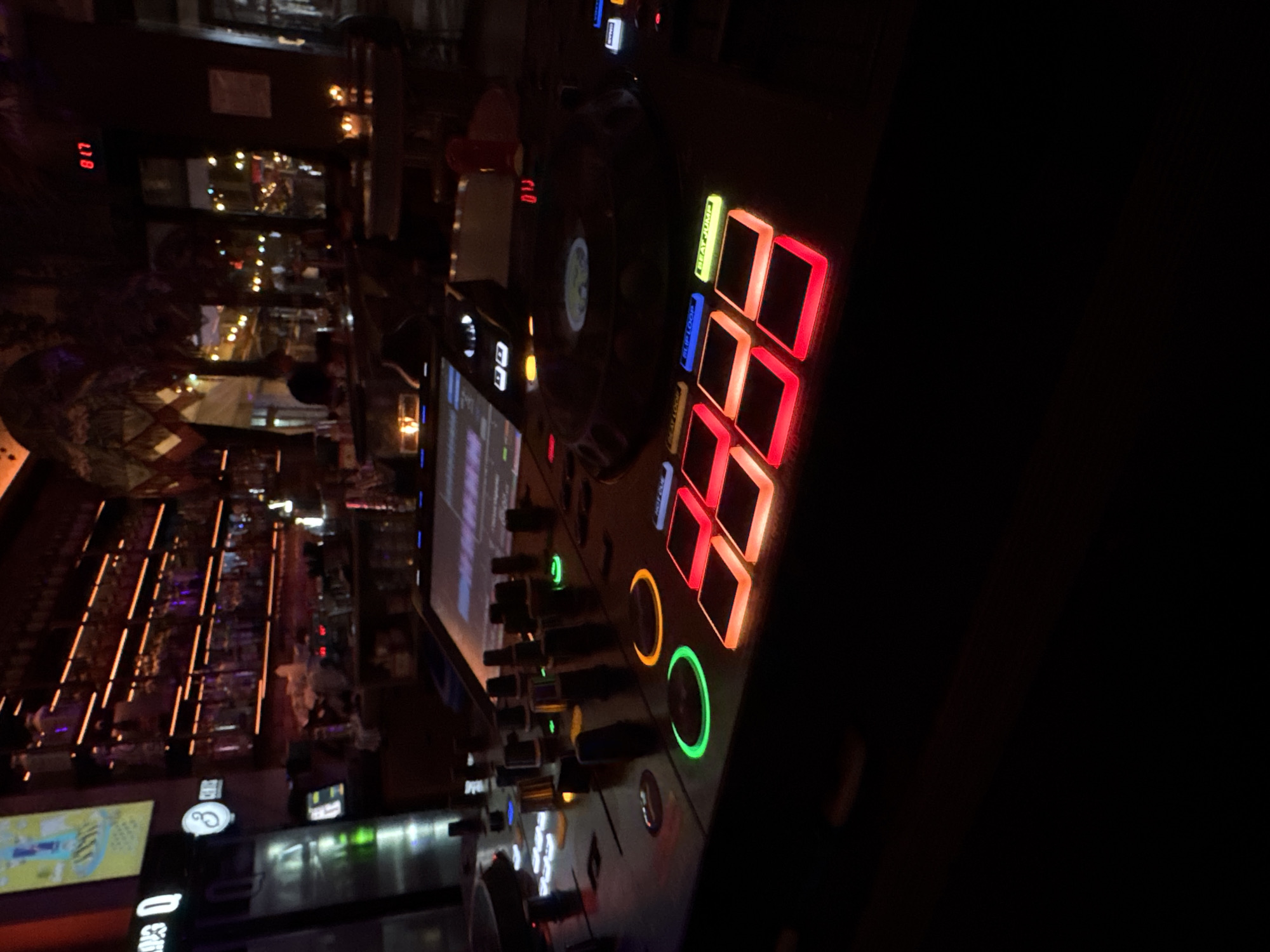 The image shows a DJ setup in what appears to be a bar or nightclub in Plovdiv, Bulgaria. The DJ console is prominently displayed with illuminated buttons and controls, some in red and green. The surrounding area is dimly lit but reveals a view of the bar area with tables, seating, and lighting fixtures. Several people are visible in the background, and the overall atmosphere suggests a vibrant nightlife setting.