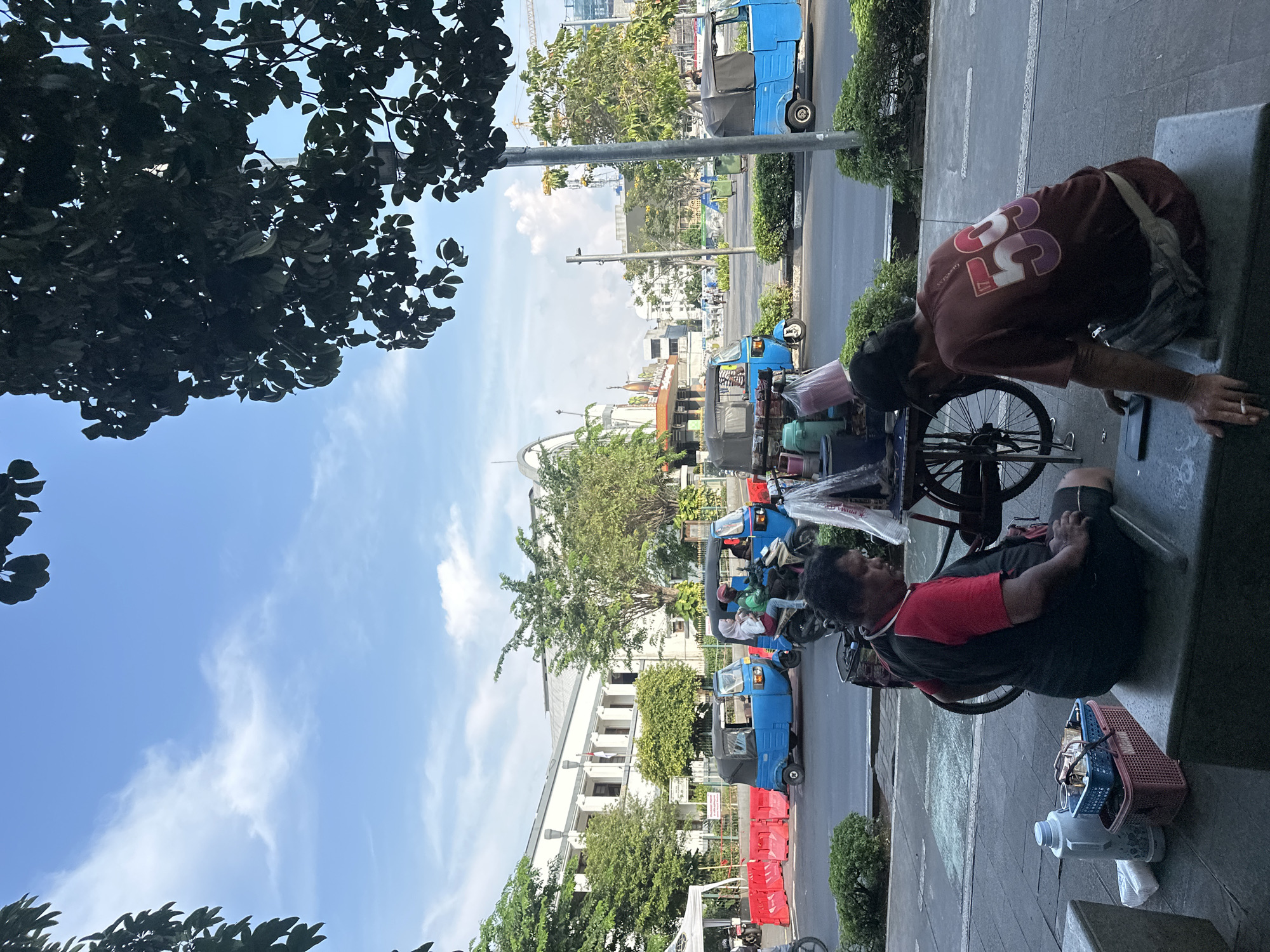 A scene in West Jakarta, Indonesia, shows a street view with pedestrians and auto rickshaws, commonly known as "bajaj." The image appears to be taken from a somewhat elevated position, looking out over the street.
In the foreground, two individuals are near a concrete structure, possibly a bench or low wall. One person is seated, wearing a red and black shirt, and has a basket and water bottles nearby. The other person, wearing a maroon shirt with the number 33 on the back, leans over the bench. There is a bicycle leaning against the bench.
The background features several blue auto rickshaws parked along the side of the road. Some of these vehicles have passengers. A row of red and orange traffic barriers is visible on the road's edge. In the distance, buildings, trees, and a partly cloudy sky can be seen. A construction crane is visible in the distance. The overall impression is a typical urban street scene in West Jakarta.