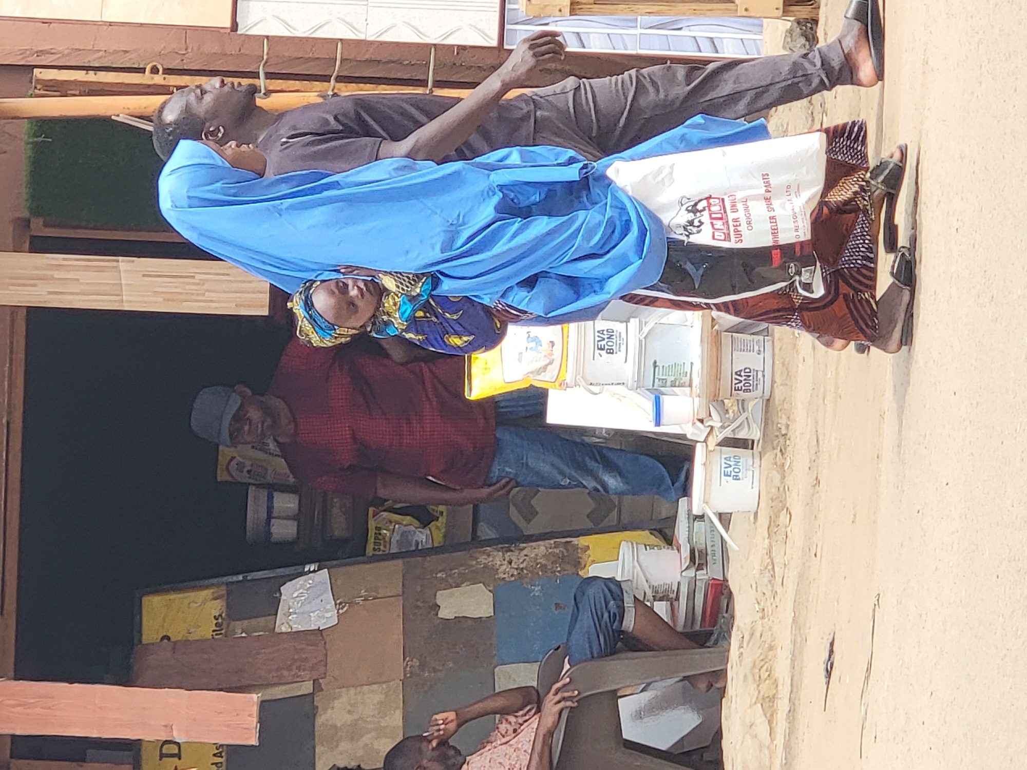 A medium, eye-level, angled shot captures a public scene in Maiburiji, Nigeria, highlighting three visible men and a woman carrying a load.
Closest to the foreground is a sandy-colored pavement. To the left is an array of construction materials and two men. One man is seated, leaning back in a plastic chair, and is smoking. Further back is a second man wearing a red shirt and a grey baseball cap. He is standing and faces right.
In the foreground is a couple carrying something heavy. A man is walking on the right edge. He's wearing dark pants, a dark short-sleeved t-shirt, and dark shoes with no socks. A woman, partially obscured behind him, is carrying a large bag. She is wearing a light blue Hijab and brown patterned shoes. The bag has a “UNICO” logo on it.