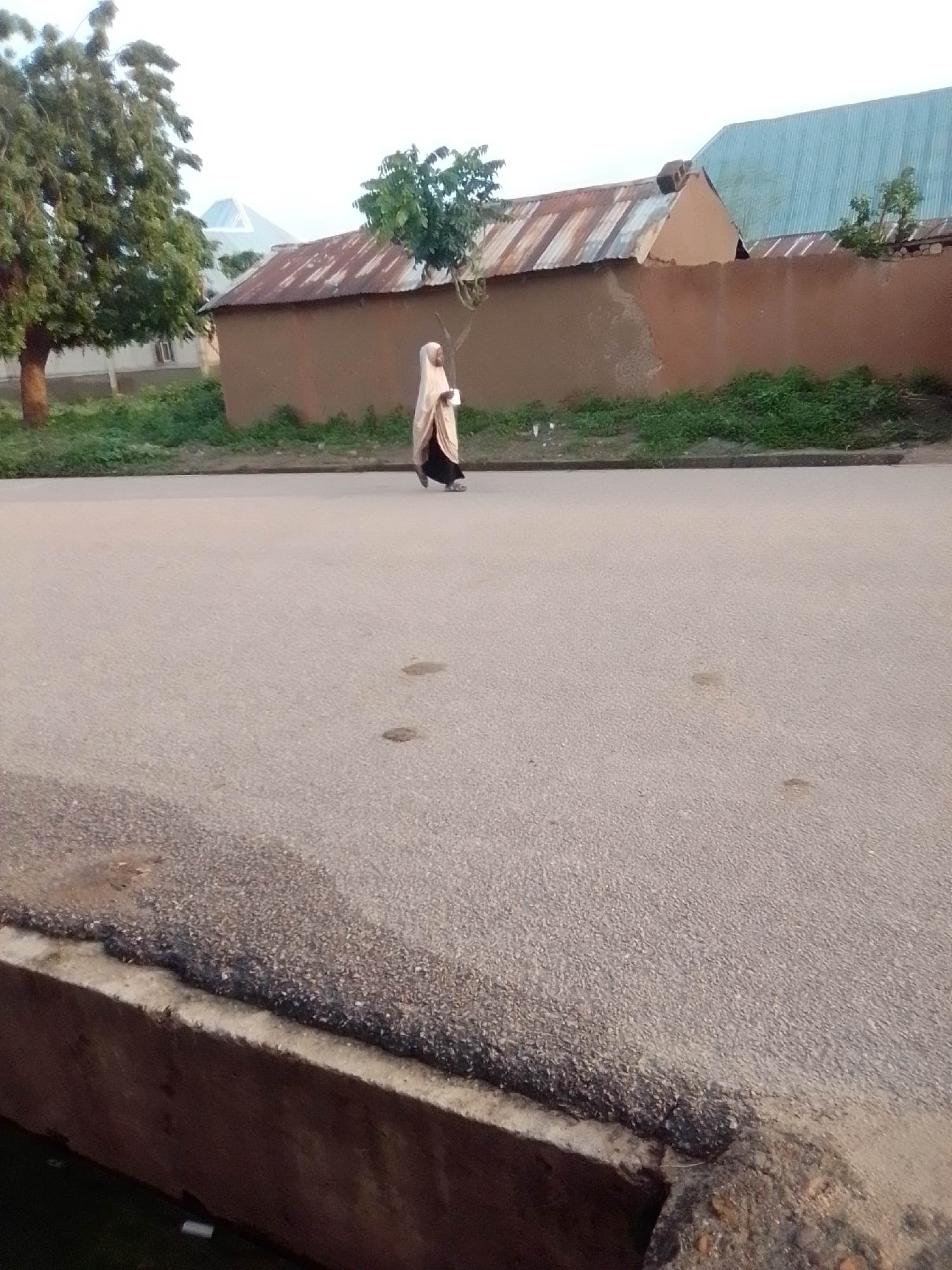 A person wearing a light-colored hijab and a dark garment is walking on a paved road. The person is positioned in the middle ground of the image, slightly to the left of center. Behind the person, there is a low-rise building with a corrugated metal roof and brown mud walls. Trees and vegetation are visible to the left and right of the building. The sky is overcast. The scene is likely situated in a residential area of Gombe, Nigeria. The road surface is asphalt with some visible patches of discoloration. There is a concrete barrier or curb in the foreground.