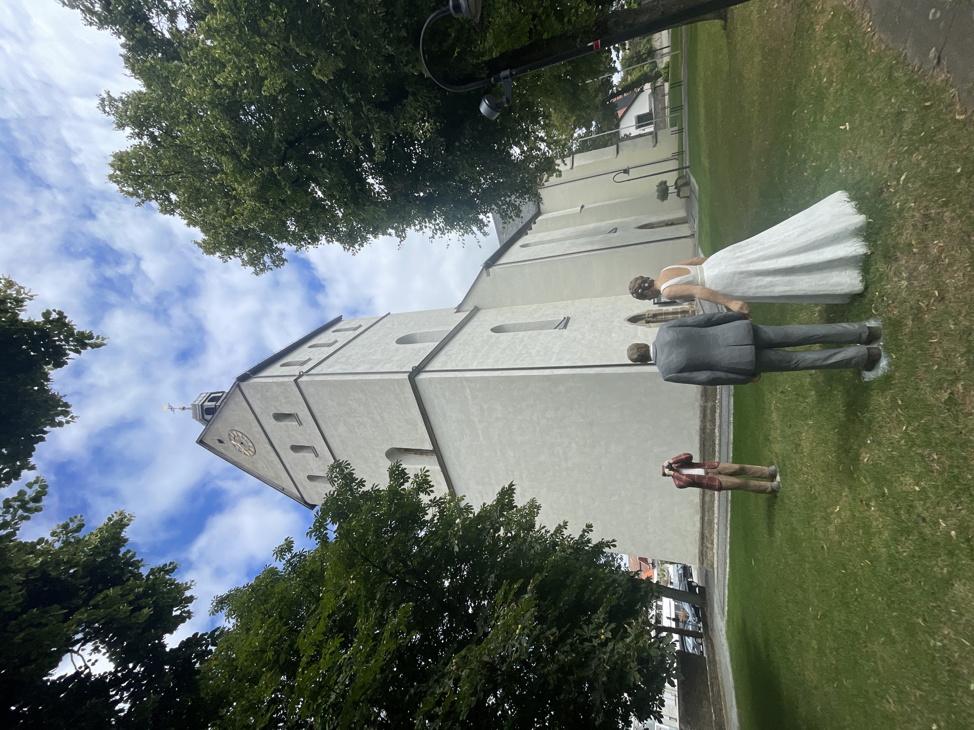 The image depicts an outdoor scene in front of a white church, identified as the St. John's Church in Salzkotten, Germany. The church features a prominent tower with a clock face and a cross on top. The foreground is dominated by a grassy lawn.

Positioned on the lawn are three life-sized human sculptures. In the center, a male sculpture dressed in a grey suit is holding hands with a female sculpture wearing a white wedding dress and her hair styled with flowers. To the left, another male sculpture is standing, wearing a red plaid jacket, brown pants, and holding what appears to be a camera to his face, as if taking a photograph.

Surrounding the church and lawn are mature green trees, with branches extending into the frame from the top left and right. The sky above is blue with scattered white clouds. In the background, beyond the church, parts of the town of Salzkotten are visible, including buildings and parked cars. A street lamp is also present to the right of the frame. The overall impression is that of a static, posed scene, likely a public art installation or a memorial.