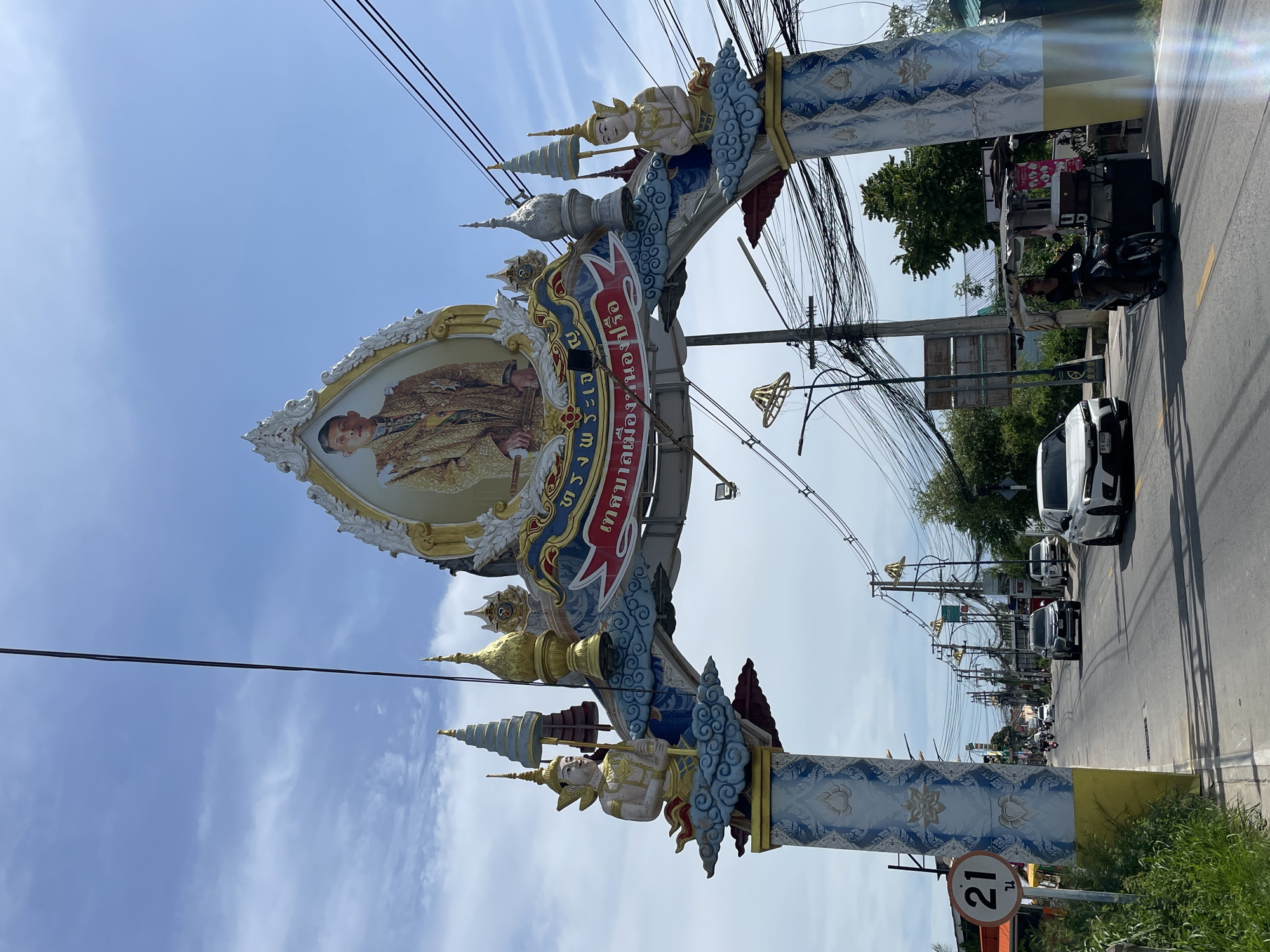 The image captures a wide view of an ornate archway in Nong Prue, Thailand, with a street scene unfolding beneath it. The archway is decorated with a large portrait of a man in formal attire, likely a royal figure, at its apex. Two angelic figures, one on each side, are perched on clouds adorning the pillars. The pillars themselves are painted with intricate blue and white patterns.
Below the archway, a two-lane asphalt road stretches into the distance, lined with trees, utility poles laden with tangled wires, and various buildings. Several vehicles are visible on the road: a white SUV, a white sedan, a motorcycle, and a small cart attached to a motorcycle. A speed limit sign indicating "21" is present on the left side of the road. The sky above is a clear blue with scattered white clouds. The overall impression is a vibrant and busy urban environment in Thailand.