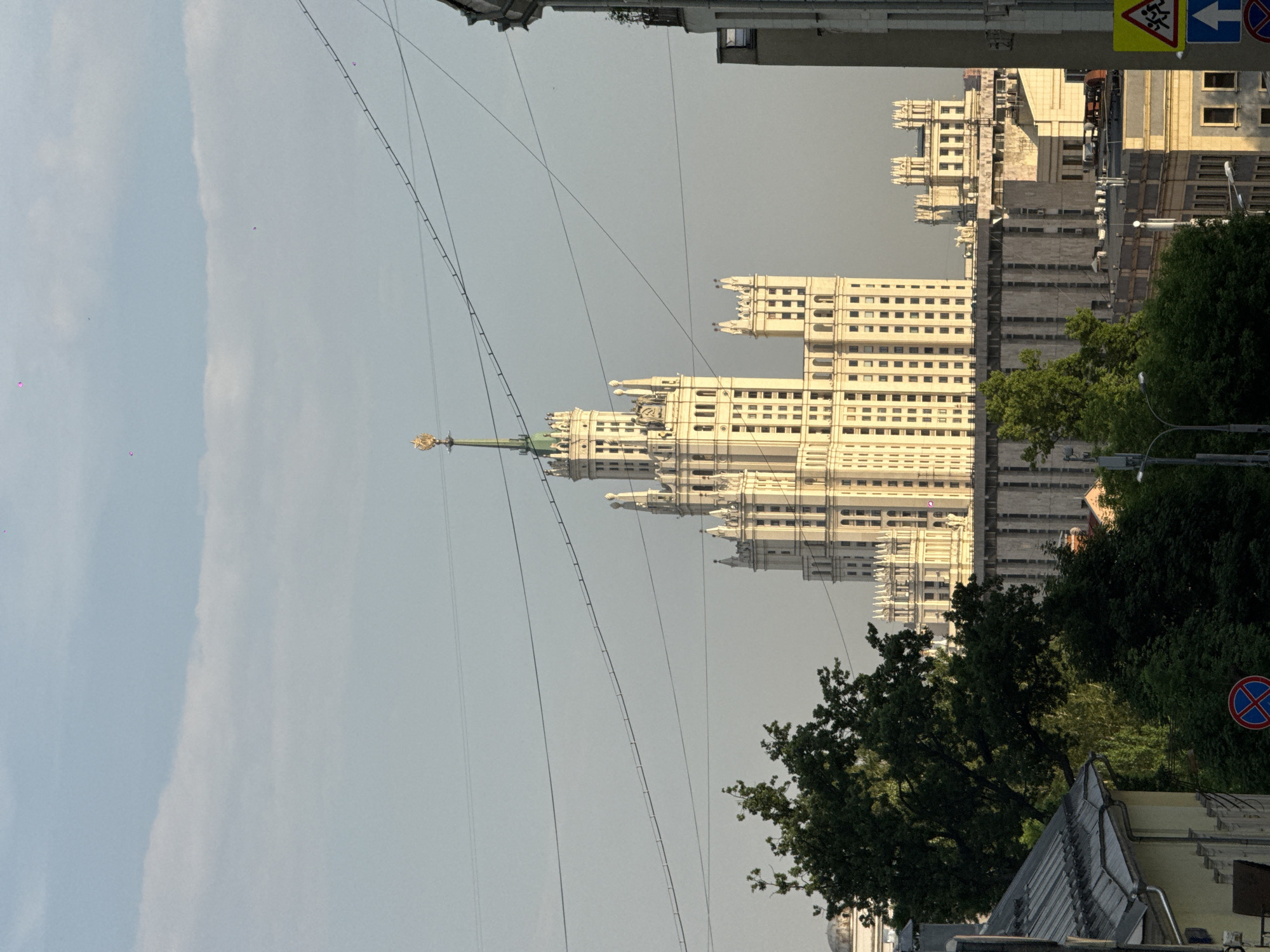 A slightly rotated, daytime, street-level view of the Kotelnicheskaya Embankment Building in Moscow, Russia. The skyscraper is prominently featured in the center, composed of multiple tiers with numerous windows. Above its highest point is a golden spire. The sky is overcast. High-tension cables run diagonally across the image. To the right are trees and sections of buildings, including a street sign. To the left is sky.