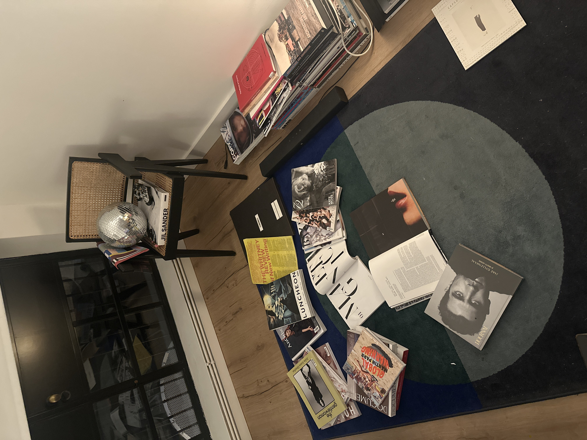 A slightly elevated, interior shot reveals a collection of books, magazines, and objects spread across a geometrically patterned rug on a wooden floor, next to a black-framed glass door and adjacent to a collection of books leaning against a wall.
To the left, a disco ball and a book titled "JIL SANDER" rest on a chair with a wicker seat. The nearby black-framed glass door reflects the interior.
On the wooden floor, a geometrically patterned rug features circles and squares in shades of black, grey, green, and blue. Scattered across the rug are numerous books and magazines. Titles visible include "LUNCHEON," "THE WHITNEY REVIEW of New Writing," "THE GENTLEWOMAN," "GLOBAL MARTIN PARR," "ARCHITECTURE AND ANARCHISM," and "DAZED."
A black soundbar rests between the rug and a collection of books leaning against the wall. These books include "COPY," "ARCHITECTURE AND ANARCHISM," and a red book with visible text. A white cable is visible near this collection.
The scene appears to be in a residential or studio space, possibly in London, United Kingdom, given the style and content of the books and magazines. The lighting is soft, indicating a diffused light source. There are no people visible in the shot. The layout suggests a disorganized workspace or a temporary arrangement of the items.