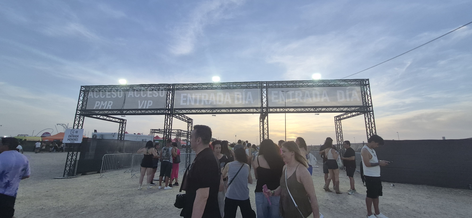 The image depicts an outdoor scene at what appears to be an event entrance under a cloudy sky. A large metal truss structure spans across the frame, adorned with banners. One banner reads "ACCESO ACCESO PMR VIP", indicating access for people with reduced mobility and VIPs. Another banner, repeated multiple times, says "ENTRADA DÍA", which translates to "Day Entry". To the left, a smaller sign displays a "18" symbol with "ENTRADA MENORES" below it, signifying that those under 18 are not permitted entry.
A crowd of people is visible, gathered and queuing near the entrance. They are dressed in casual summer attire. In the foreground, a man wearing a black t-shirt and a man in a purple t-shirt with a palm tree print are prominent. People are in various states of waiting, some looking at their phones, others conversing. The ground is a sandy or gravelly surface, typical of outdoor event venues. In the background, tents and structures can be seen, suggesting a festival or concert. The time of day appears to be late afternoon or early evening, with the sun setting or having recently set, casting a warm glow on the horizon visible through the truss. The scene is likely situated in Arganda del Rey, Spain, given the context provided.
