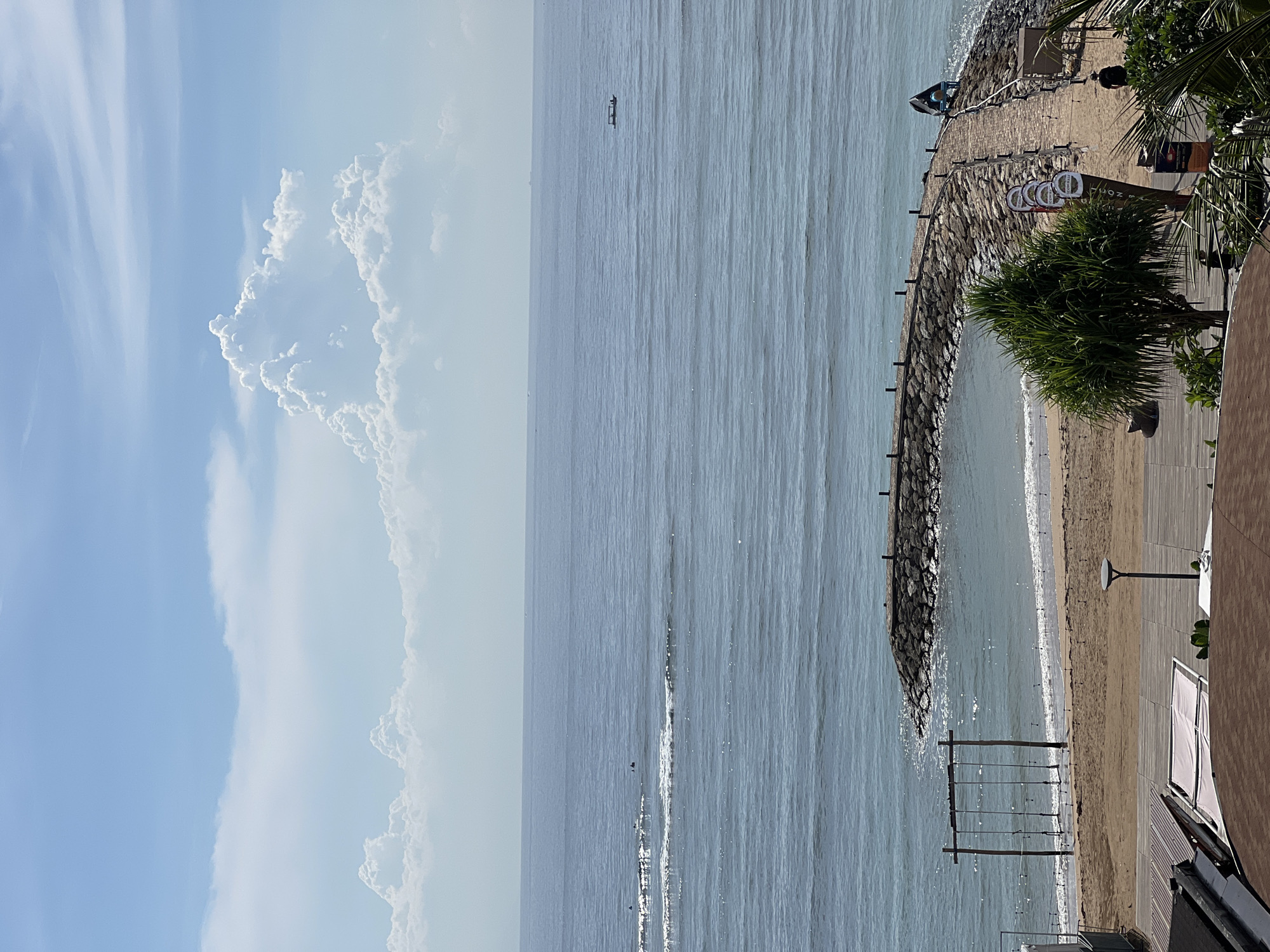 The media file presents a bright, panoramic view of a coastal scene in Denpasar, Indonesia, dominated by the ocean, sky, and a developed shoreline. The image is oriented vertically but depicts a horizontal landscape.
The upper portion of the image shows a clear blue sky dotted with scattered white clouds. To the right, a large, fluffy cumulus cloud formation is prominent, while the left features more wispy, high-altitude clouds. Bright sunlight suggests a clear day.
The central and largest part of the image is the expansive, calm ocean, appearing a light blue-grey. The water's surface is lightly rippled, reflecting sunlight in various glints. In the distance, a small, dark object, likely a kayak or a small boat, is visible on the horizon. Closer to the shore, gentle waves break, creating a thin line of white foam.
The right side of the image features the shoreline. A stone breakwater or jetty curves along the coast, separating the open water from a sandy beach. Further inland from the beach, a wooden boardwalk or paved path runs parallel to the shore. Numerous amenities are present along this area: several beach chairs with white coverings are arranged on the boardwalk, and lush green palm trees and other tropical foliage are scattered throughout. A tall street lamp stands on the boardwalk. In the immediate foreground, part of a building with a brown facade and a white awning is visible, likely a resort or restaurant structure. A wooden swing set structure, consisting of four vertical posts and horizontal bars, extends into the shallow water near the beach.
Two people are discernible in the scene: one individual in dark clothing stands near the palm trees on the boardwalk, and another person wearing a blue top is visible near the water, next to a blue object that could be a small boat or a shaded seating area.
On some of the beach chairs or nearby signs, the text "COCO" is visibly printed multiple times in a circular emblem. The overall scene conveys a serene and inviting atmosphere typical of a resort beach during daytime.