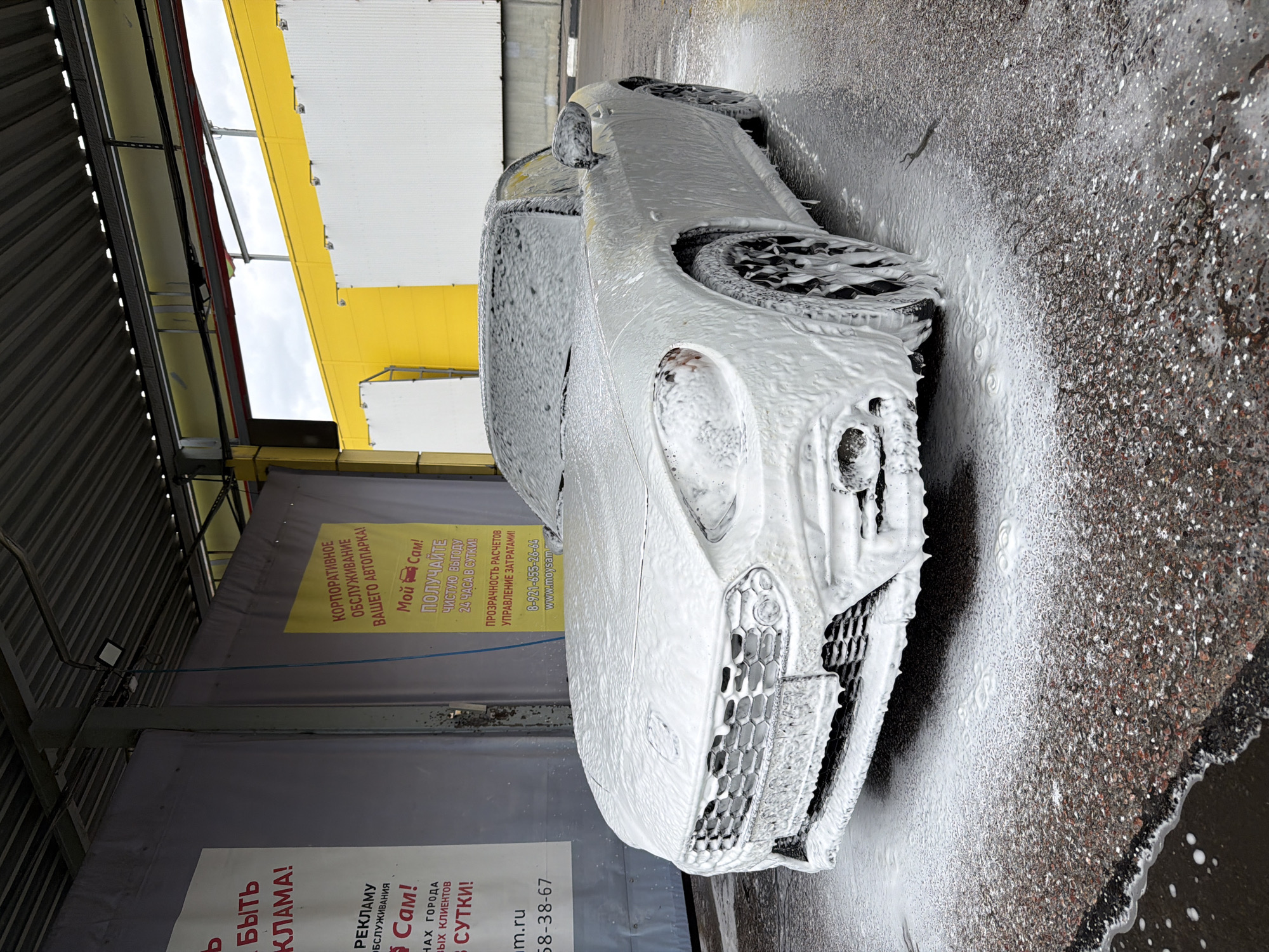 The image shows a car covered in foam at a self-service car wash. The location is identified by a sign with Russian text, including "Мой Сам!" (My Sam!), a brand name, and "8-921-655-26-64," a phone number associated with Saint Petersburg, Russia. The signage promotes corporate car wash services. The car wash is located under a building overhang, and the car is surrounded by water and foam residue.