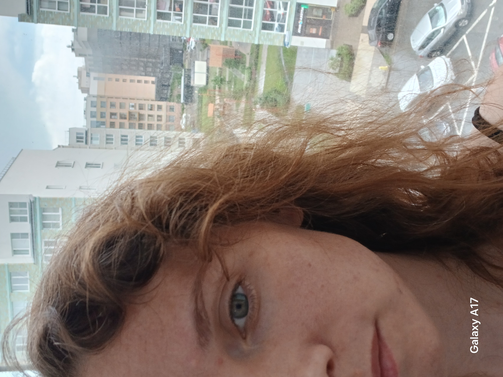 The image captures a selfie taken from an interior window looking out onto an urban landscape, rotated 90 degrees clockwise. In the immediate foreground, filling a significant portion of the frame, is the right side of a person's face, featuring wavy, reddish-brown hair cascading around their temple and cheek. A light blue-green eye is visible, along with a portion of the nose and mouth. The skin shows some freckling.
Through the window, an urban scene unfolds under an overcast, daytime sky, suggesting a muted light condition, possibly after rain or during a drizzle, as some surfaces appear wet. Several multi-story buildings, varying in height and color (white, beige, and greenish facades), are prominent, characterized by numerous rectangular windows. Below the buildings, a paved area, likely a street or parking lot, contains several parked cars, including white, black, and red vehicles. Patches of green landscaping with trees and bushes are visible between the structures. The perspective gives the impression of being several stories high.
A watermark with the text "Galaxy A17" is present in the lower right corner of the image (when viewed in its original upright orientation). Partially visible text, "PLEMIAHT," is seen on a sign on one of the background buildings. The overall setting is an urban residential or mixed-use area, consistent with the provided location context of Yekaterinburg, Russia. The person's expression is neutral, and no specific activity other than taking a selfie is apparent.