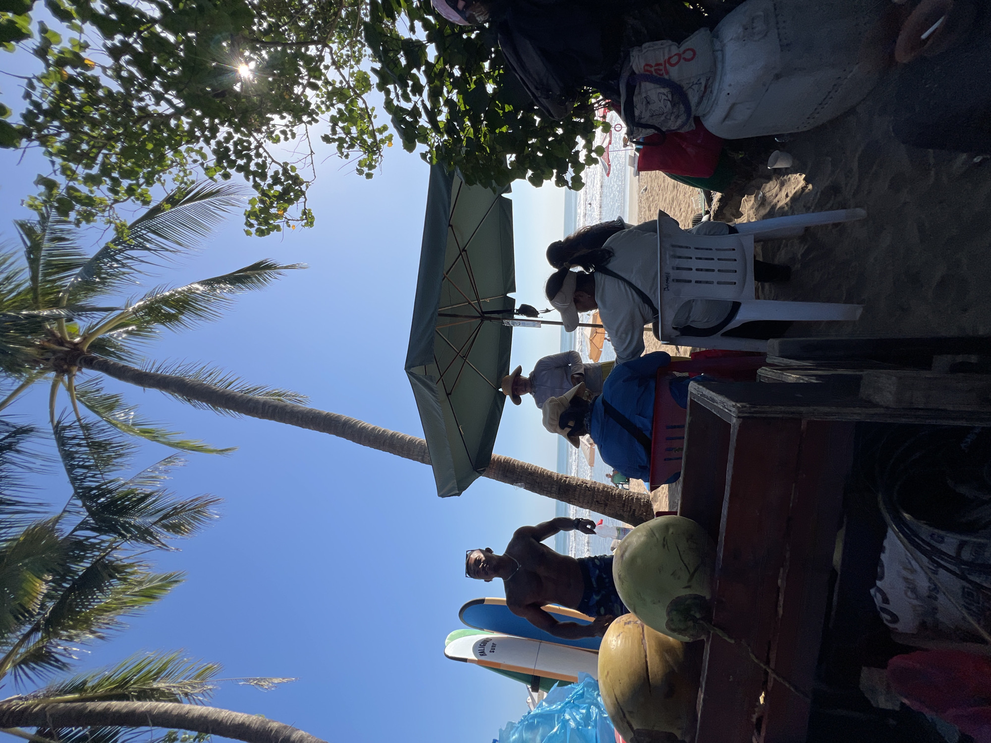The image depicts a lively scene on a beach in Kuta, Indonesia, likely a popular tourist spot. In the foreground, a wooden cart is filled with two large coconuts, suggesting a vendor selling refreshments. To the left, several surfboards are propped upright, indicating proximity to surf shops or rentals.
A shirtless man with dark skin, wearing patterned swim trunks and a necklace, stands prominently in the center, appearing to be a vendor or a local. He holds a small bottle in his right hand. Behind him, a large green umbrella provides shade over a group of people.
Several individuals are seated on chairs beneath the umbrella. One person, wearing a blue jacket and a hat, is facing away from the camera. Another person, in a white shirt and a cap, is also seated. A third person, wearing a hat and a light-colored shirt, stands slightly behind them.
The beach extends to the background, with gentle waves visible on the ocean. The sky is a clear blue, with bright sunlight filtering through palm trees and lush green foliage on the right side of the frame. The overall atmosphere appears relaxed and typical of a tropical beach setting.