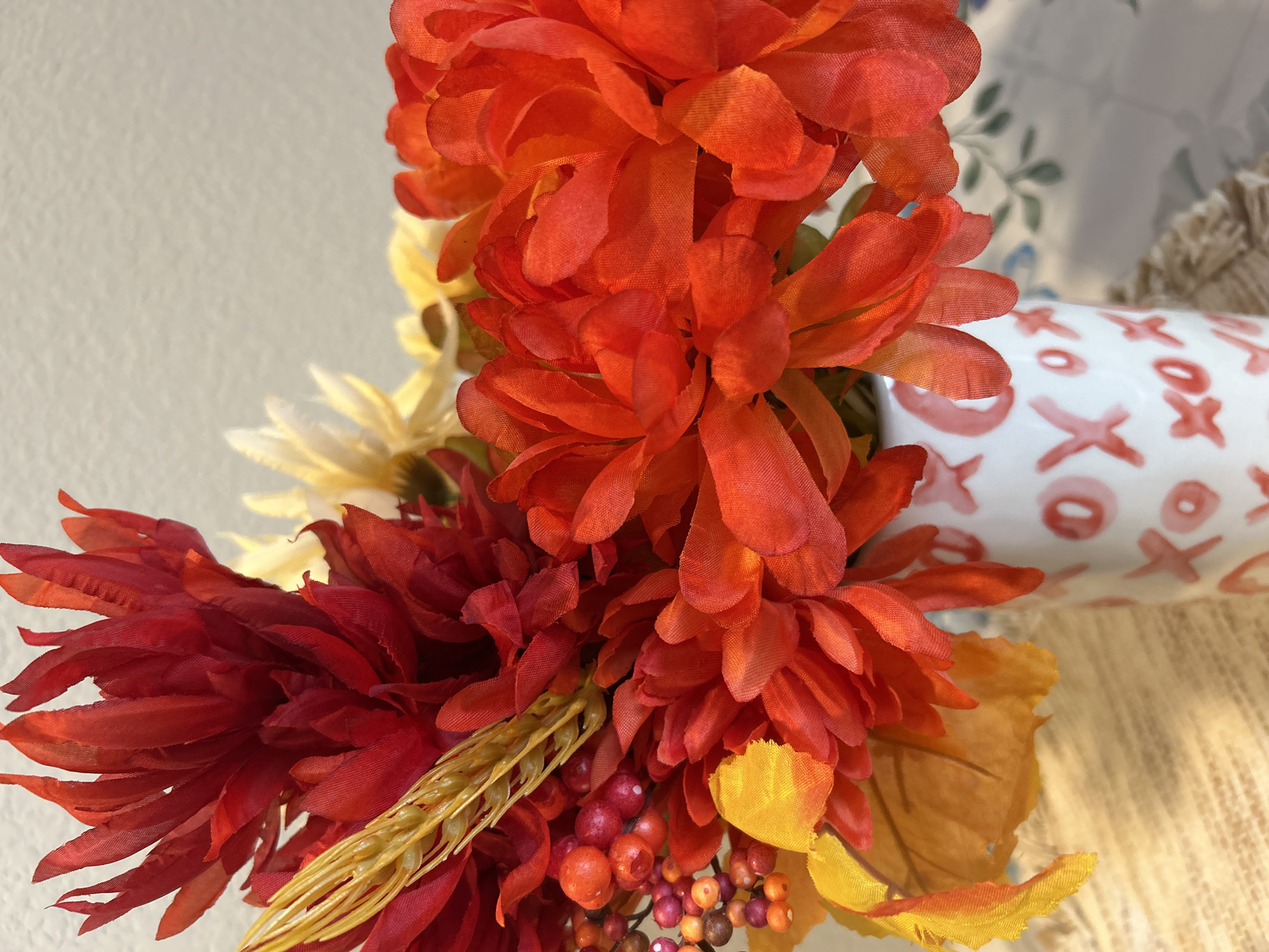 A close-up shot focuses on a vase filled with artificial flowers, predominantly in shades of red and orange, with accents of yellow and some berries. The vase itself is white and decorated with a repeating pattern of pink "X" and "O" shapes. The background is a plain, textured, light-colored wall, and the foreground shows a woven placemat. The scene appears to be indoors, likely in a home setting. No specific location in Colorado Springs, United States of America, can be determined from the visual information provided.